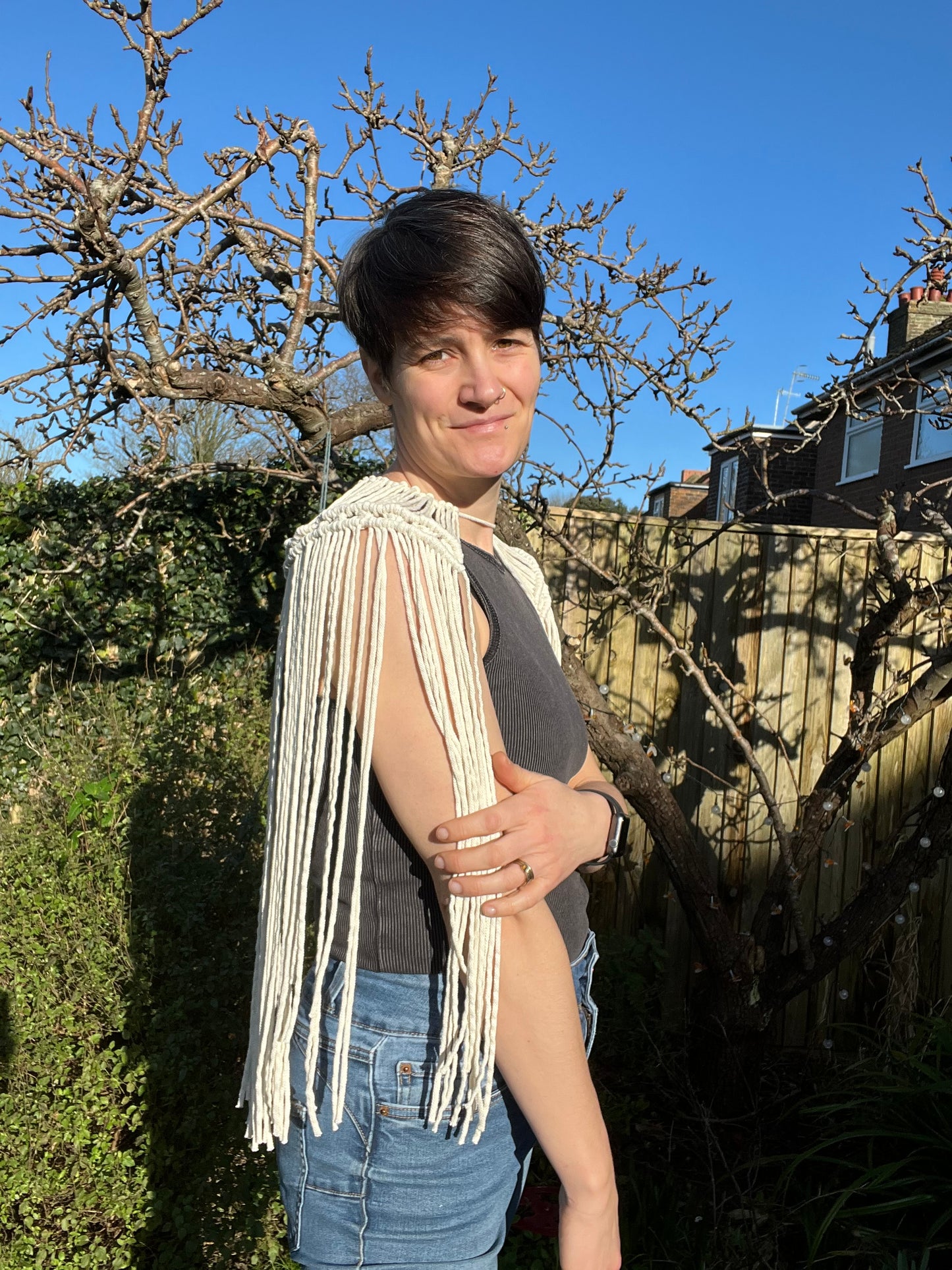 A female stands outside on a sunny day wearing a black vest, blue jeans and a handmade macrame shoulder accessory epaulette made with natural recycled cotton. She is standing side on to the camera, with one arm across her body.