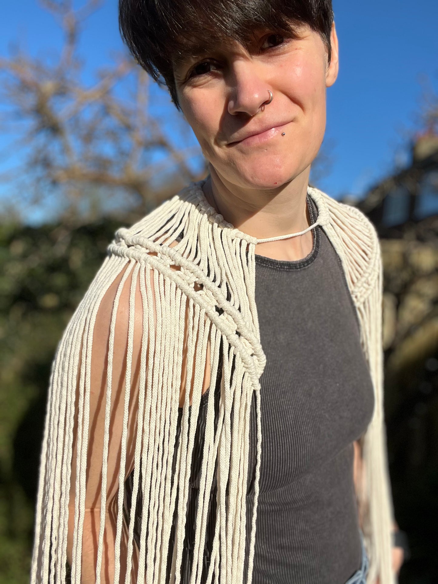 A female stands outside on a sunny day wearing a black vest and a handmade macrame shoulder accessory epaulette made with natural recycled cotton. She stands side on to the camera, leaning in and smiling.
