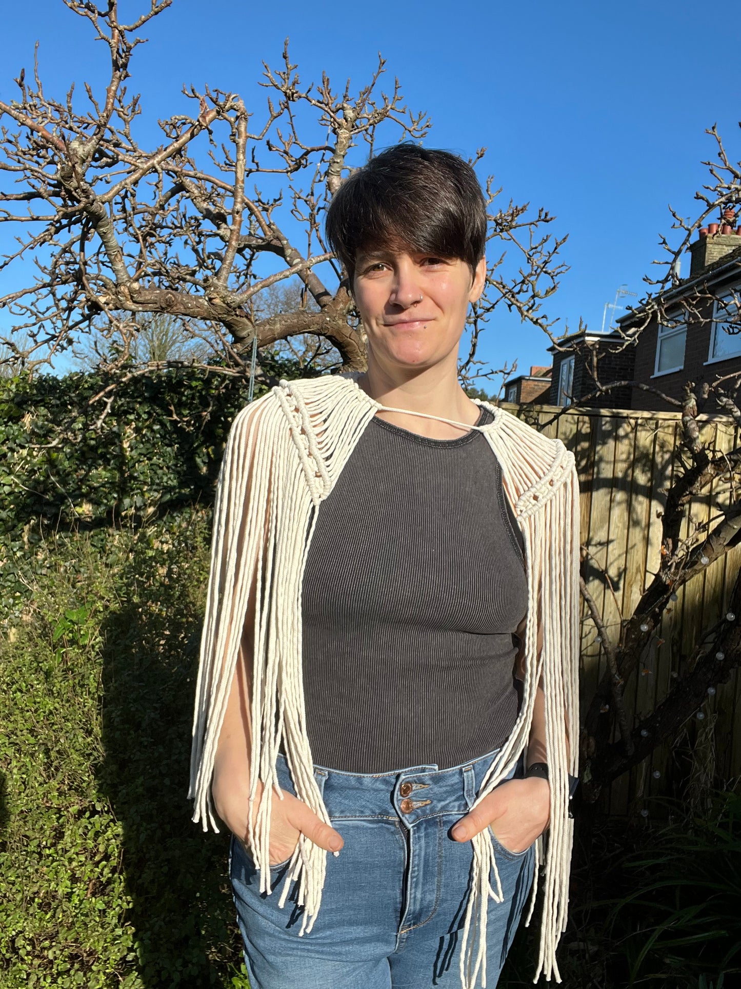 A female stands outside on a sunny day, wearing a black vest and jeans, with a handmade macrame shoulder accessory epaulette. She has her hands in the front pockets of her jeans and is looking towards the camera.