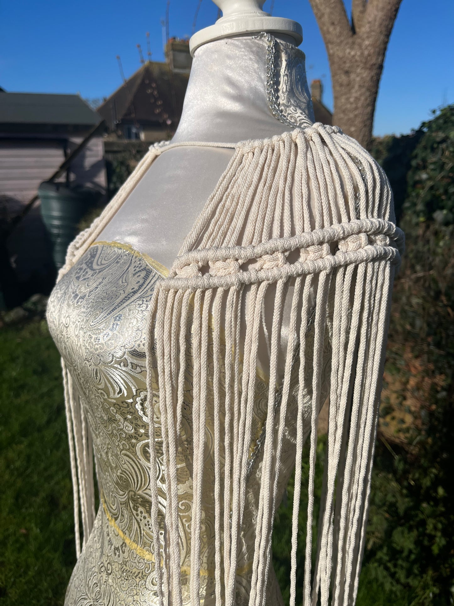 A mannequin outside in the sunshine, displays a handmade macrame shoulder accessory epaulette, made with natural recycled cotton.