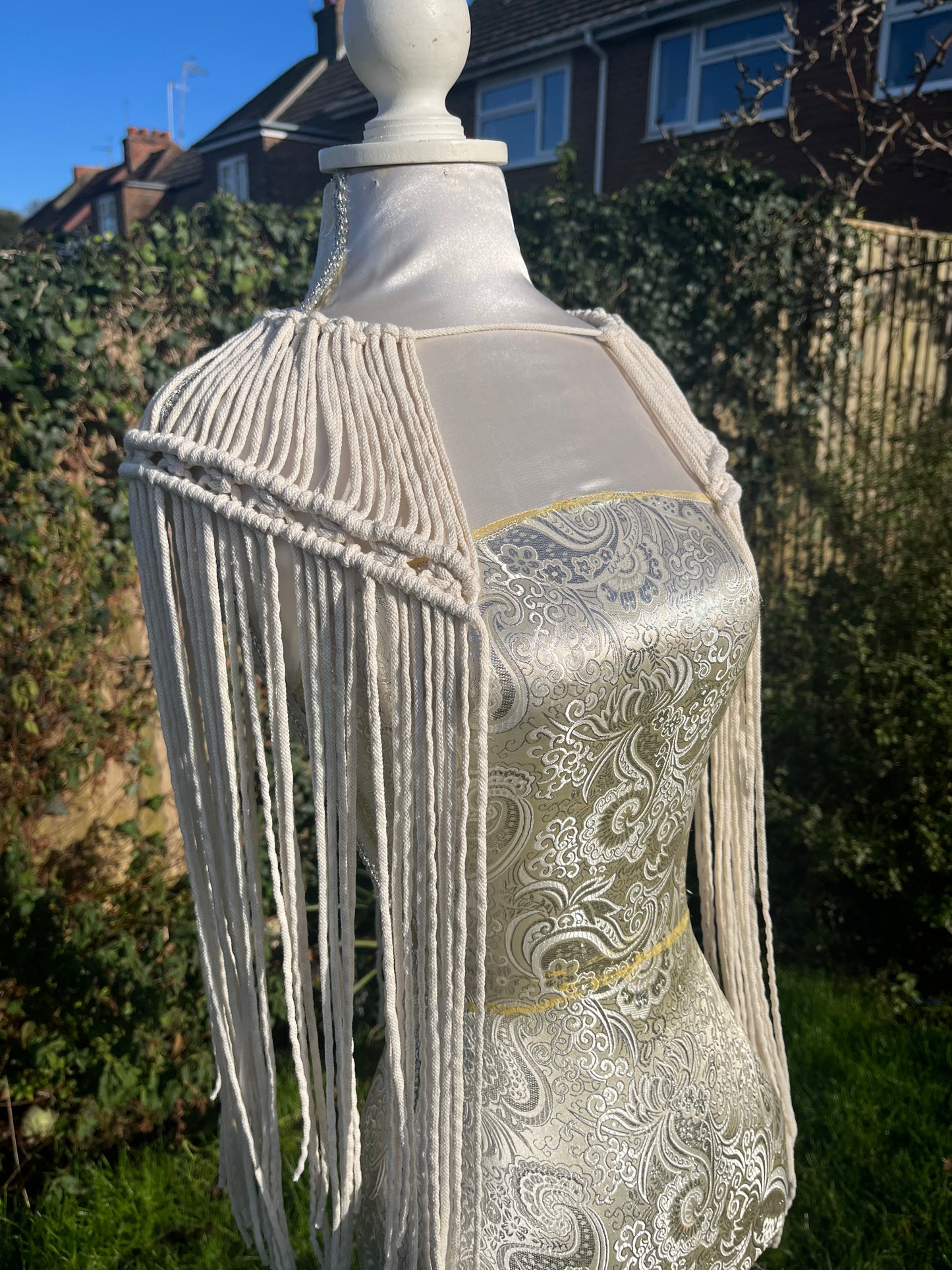 A mannequin outside in the sunshine, displays a handmade macrame shoulder accessory epaulette, made with natural recycled cotton.