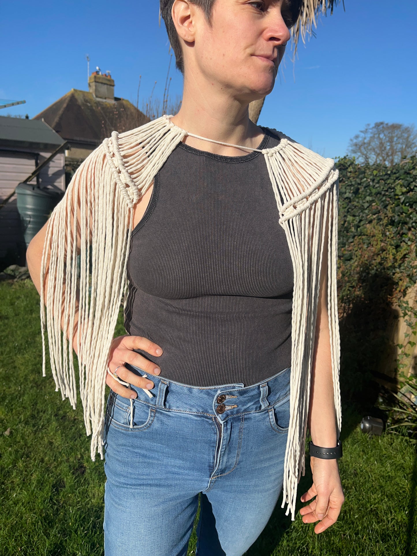 a female stands outside in the garden wearing a black vest, blue jeans and a handmade macrame shoulder accessory epaulette made with natural recycled cotton. She has one hand on her hip and is looking away from the camera.