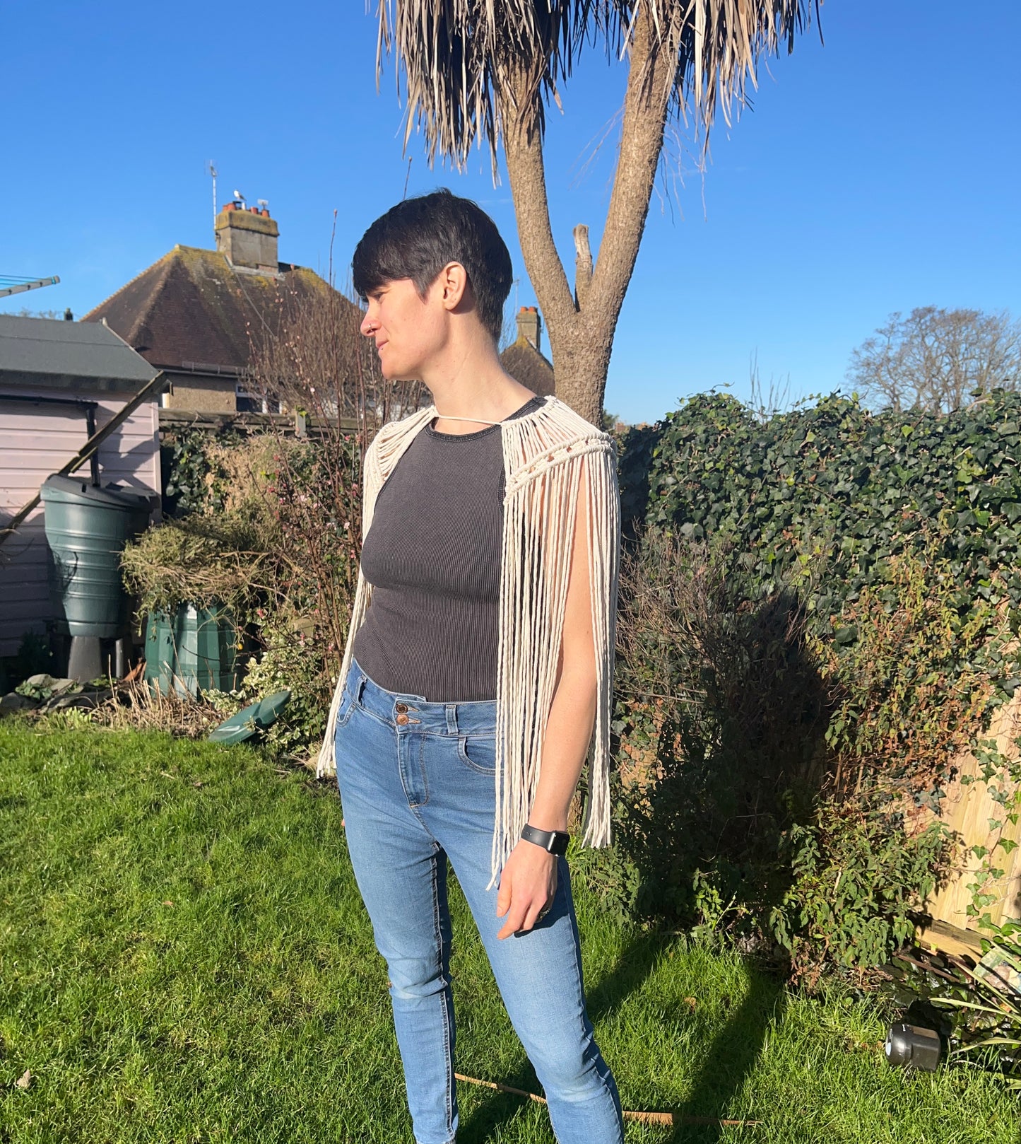 A female stands outside on a sunny day, wearing a black vest, blue jeans and a handmade macrame shoulder accessory epaulette, made with natural recycled cotton. She is looking to the side, away from the camera.