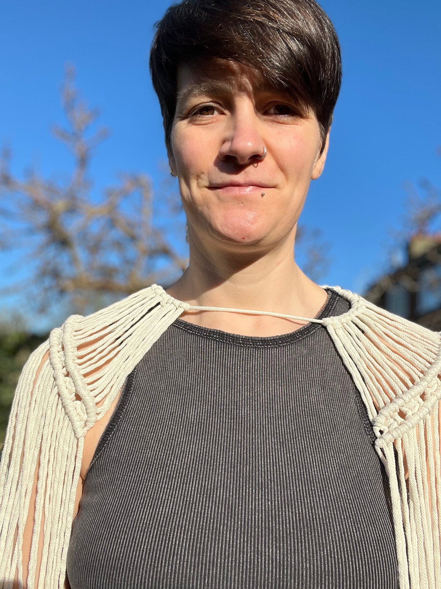 A female stands outside wearing a black vest and a handmade macrame shoulder accessory epaulette made with natural recycled cotton. She is looking directly at the camera.