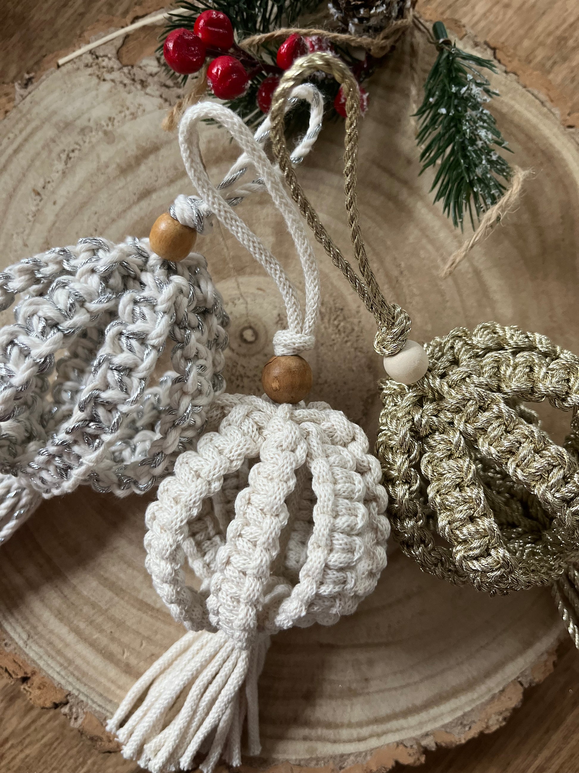 Three knotted Christmas baubles made with silver, natural and gold cotton yarns, and finished with a small wooden bead, lay beside each other on a round wooden surface.