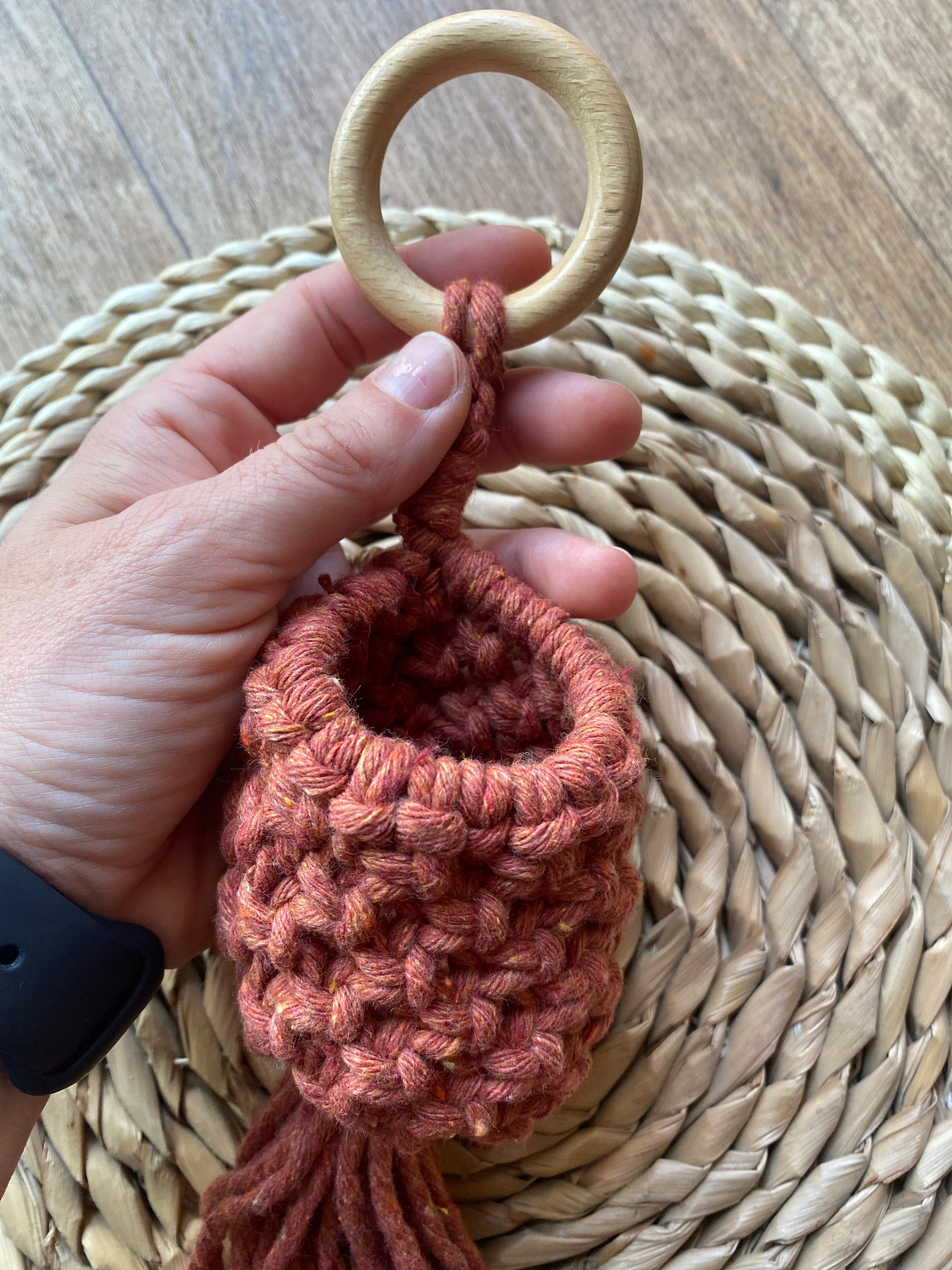 A hand holding a macrame pod hanger, made with terracotta coloured cotton and a wooden hanging ring.