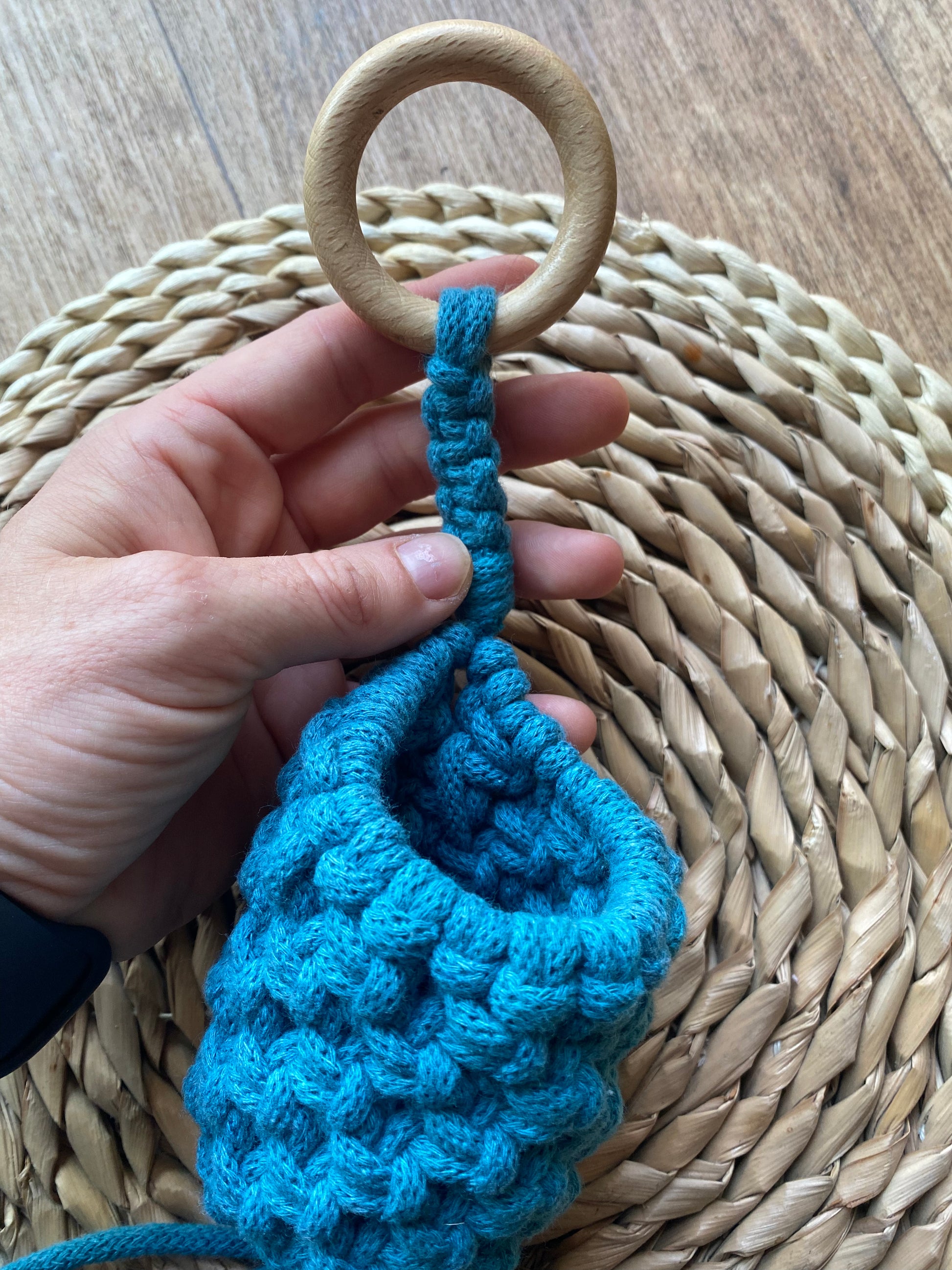 Close up of a hand holding a macrame pod hanger, made with teal blue coloured cotton and a wooden hanging ring.