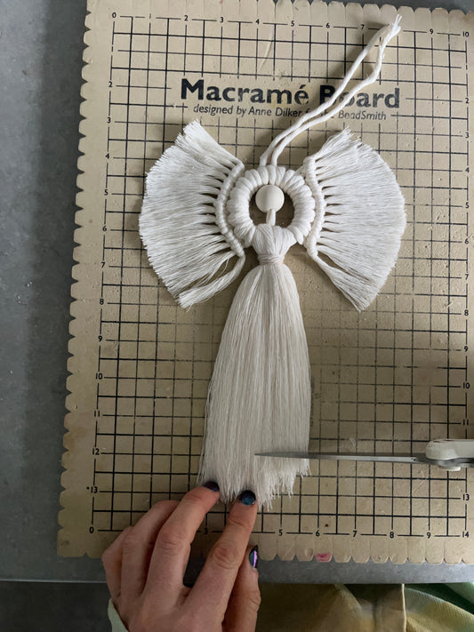 A hand about to trim the fringe on a macrame angel decoration, on a checkered board marked 'Macrame Board'.