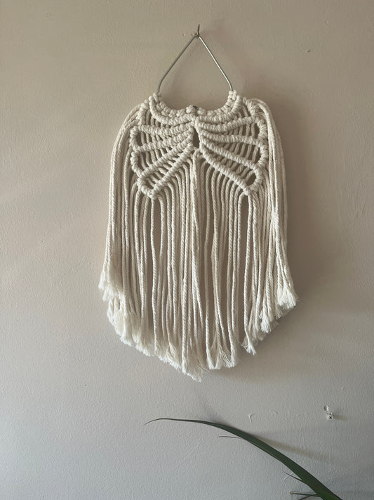 Macrame wall hanging in the shape of wings, made with natural cotton on a metal heat shaped frame, hanging against a plain background.