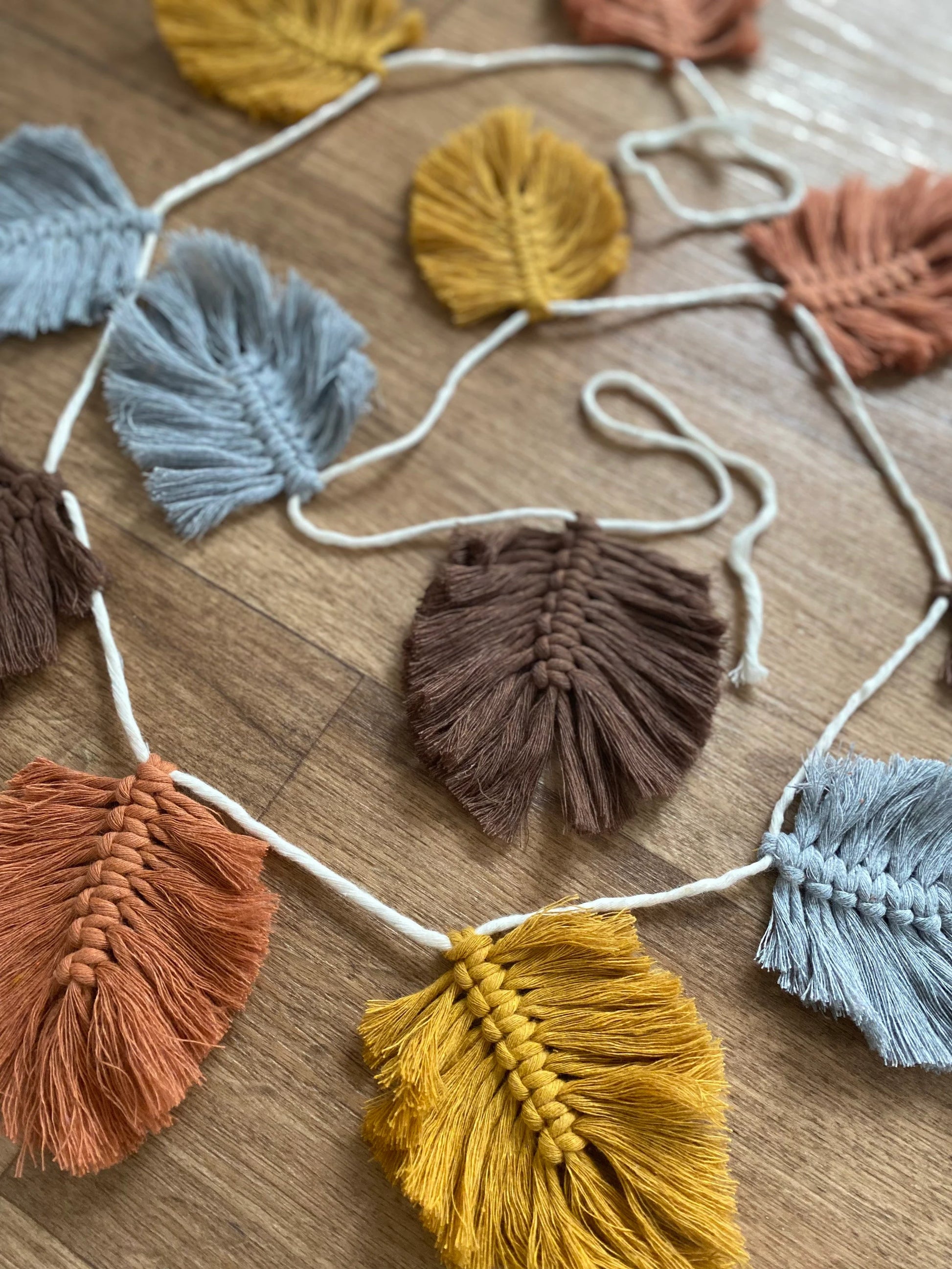 A string of 100% cotton Autumn leaf garland in various colours such as mustard yellow, grey, brown, and rust orange is laid out on a wooden surface.