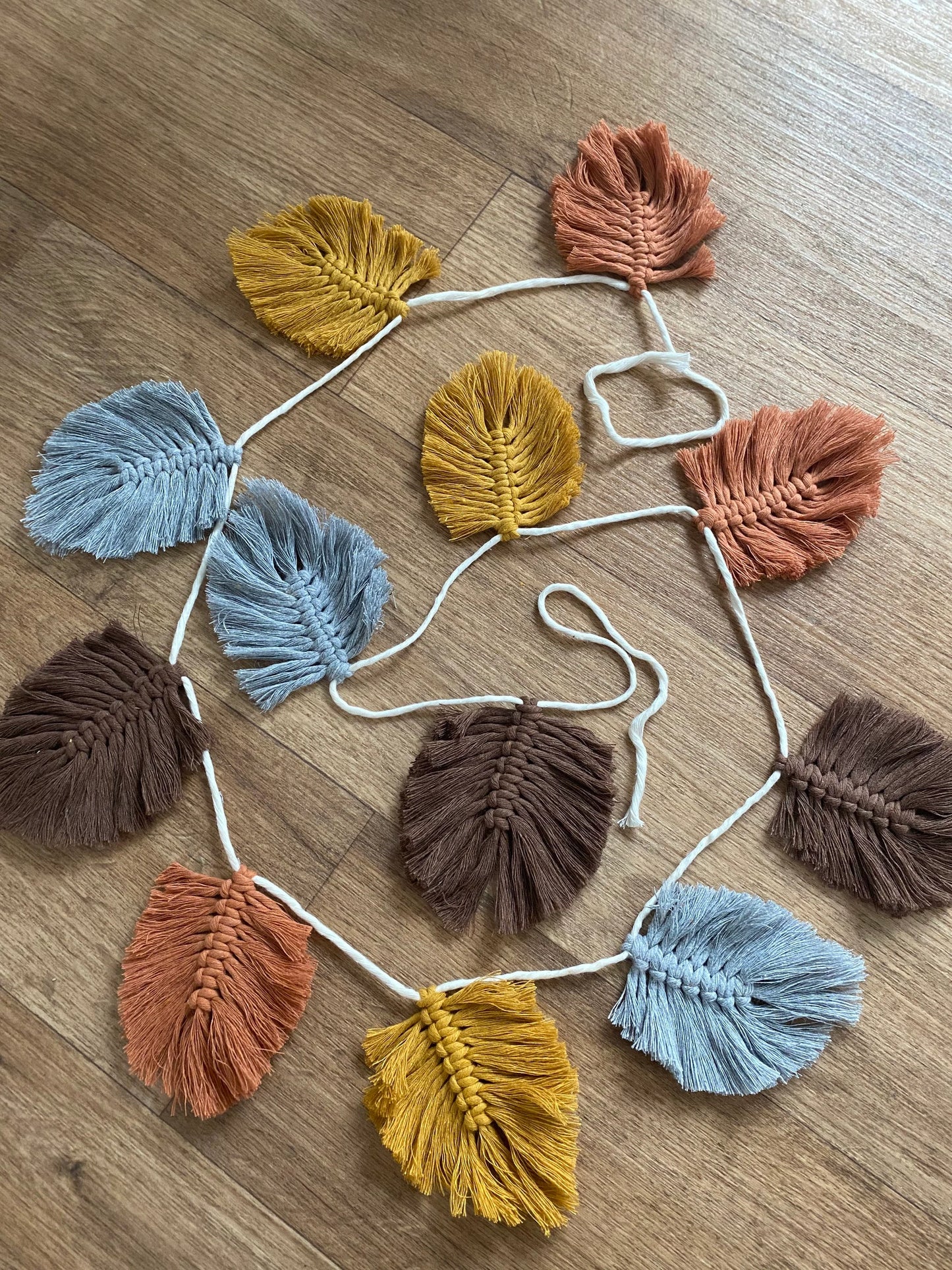 A Macra-Made-With-Love 100% cotton Autumn leaf garland featuring macramé leaves of varying colours, including grey, yellow, brown, and orange, is laid out flat on a wooden surface.