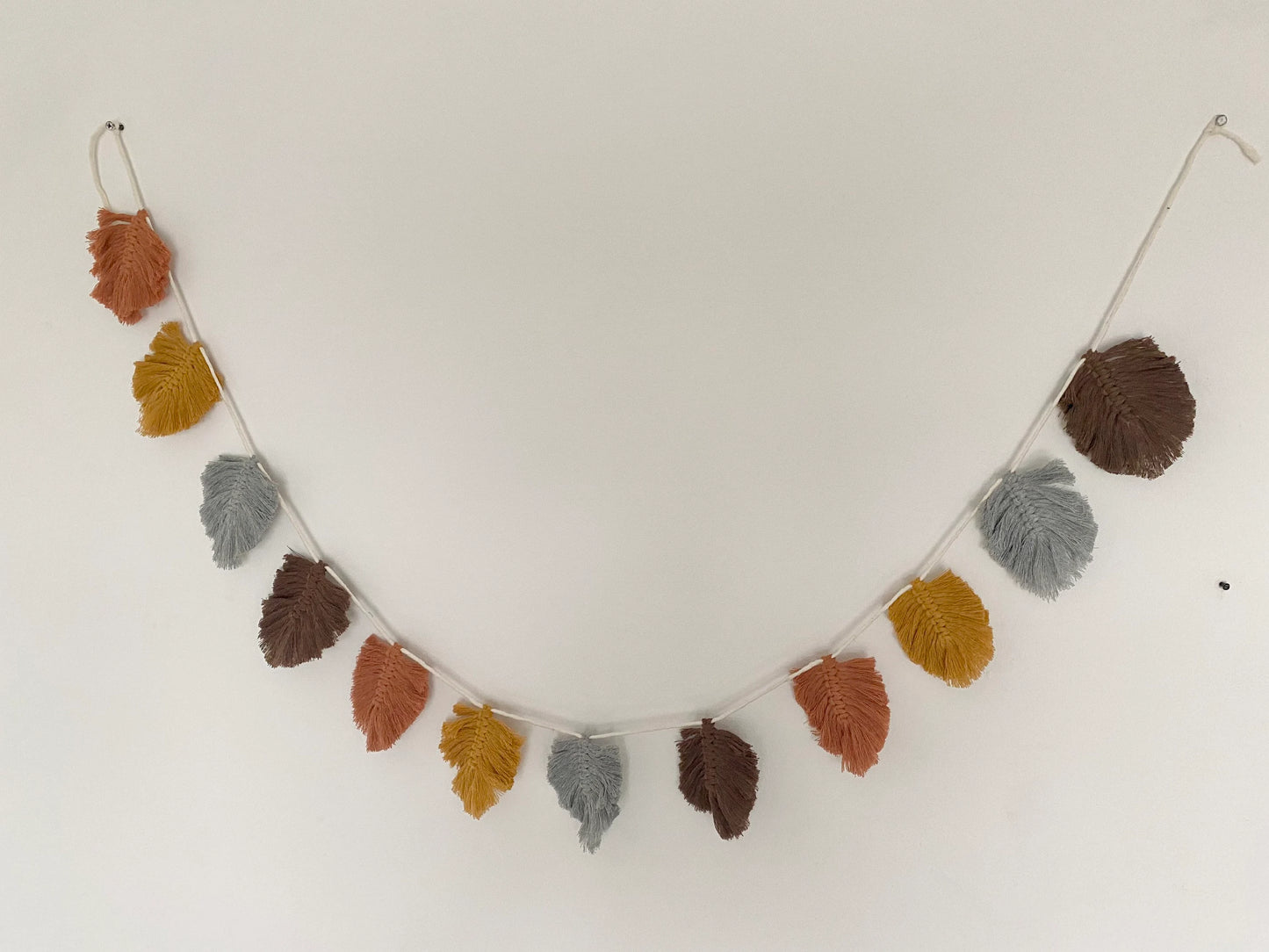 A string of decorative, macrame leaf shaped garland hangs against a white wall in autumnal colours of orange, grey, brown, and yellow.