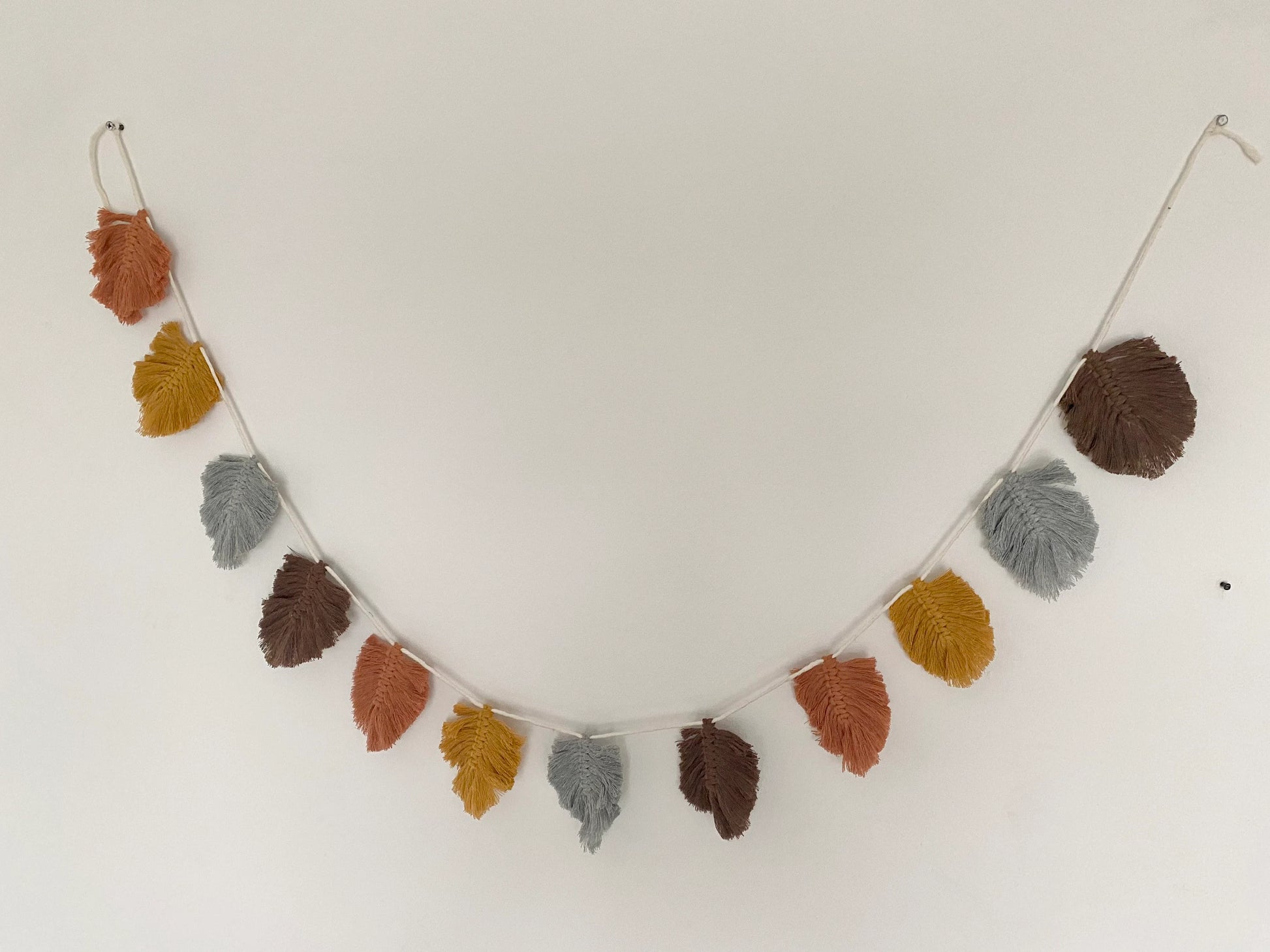 A string of decorative, macrame leaf shaped garland hangs against a white wall in autumnal colours of orange, grey, brown, and yellow.