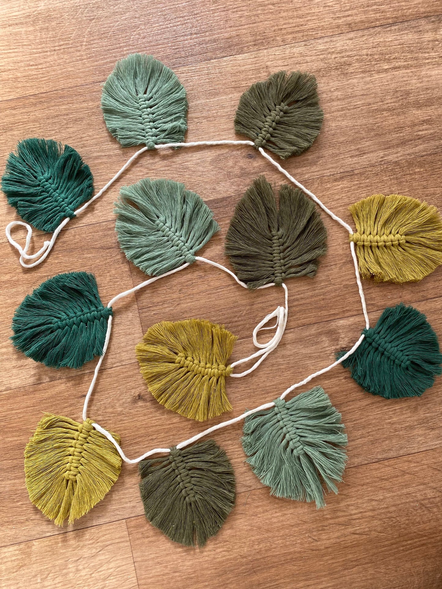 A handmade 100% cotton Autumn leaf garland from Macra-Made-With-Love in various shades of green and yellow is laid out on a wooden floor.
