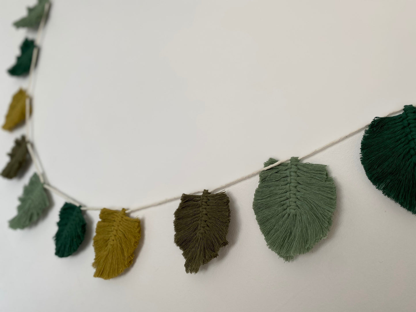 A decorative, handmade cotton garland of macrame leaves in shades of green and yellow, hanging on a string against a plain white background.