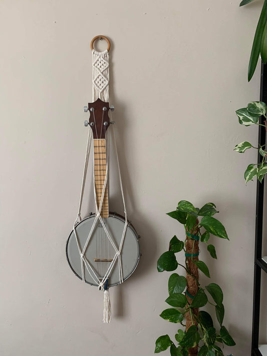 A banjolele instrument sits securely in a macrame wall mounted hanger, made with natural coloured yarn and a wooden hanging ring. Displayed against a beige painted wall, with various houseplants nearby.