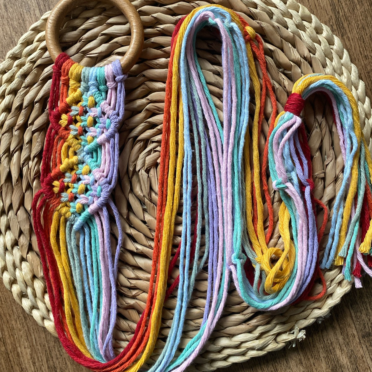 A handmade macrame banjolele wall hanger made with rainbow colours of recycled cotton on a wooden ring, laid out neatly on a woven wicker mat.