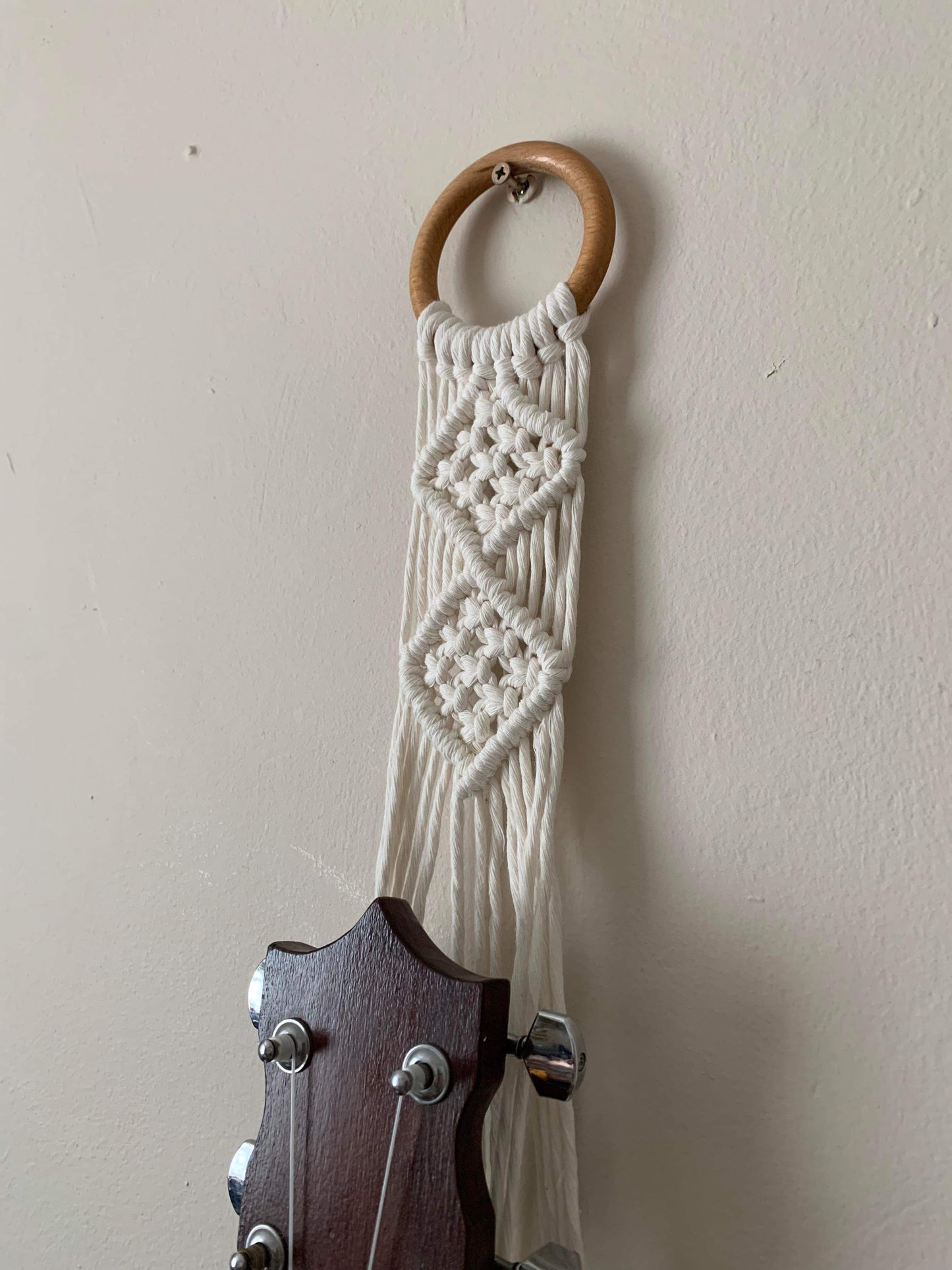 Close up image of the top section of a banjolele instrument held in a macrame wall mounted hanger, made with natural coloured yarn and a wooden hanging ring.