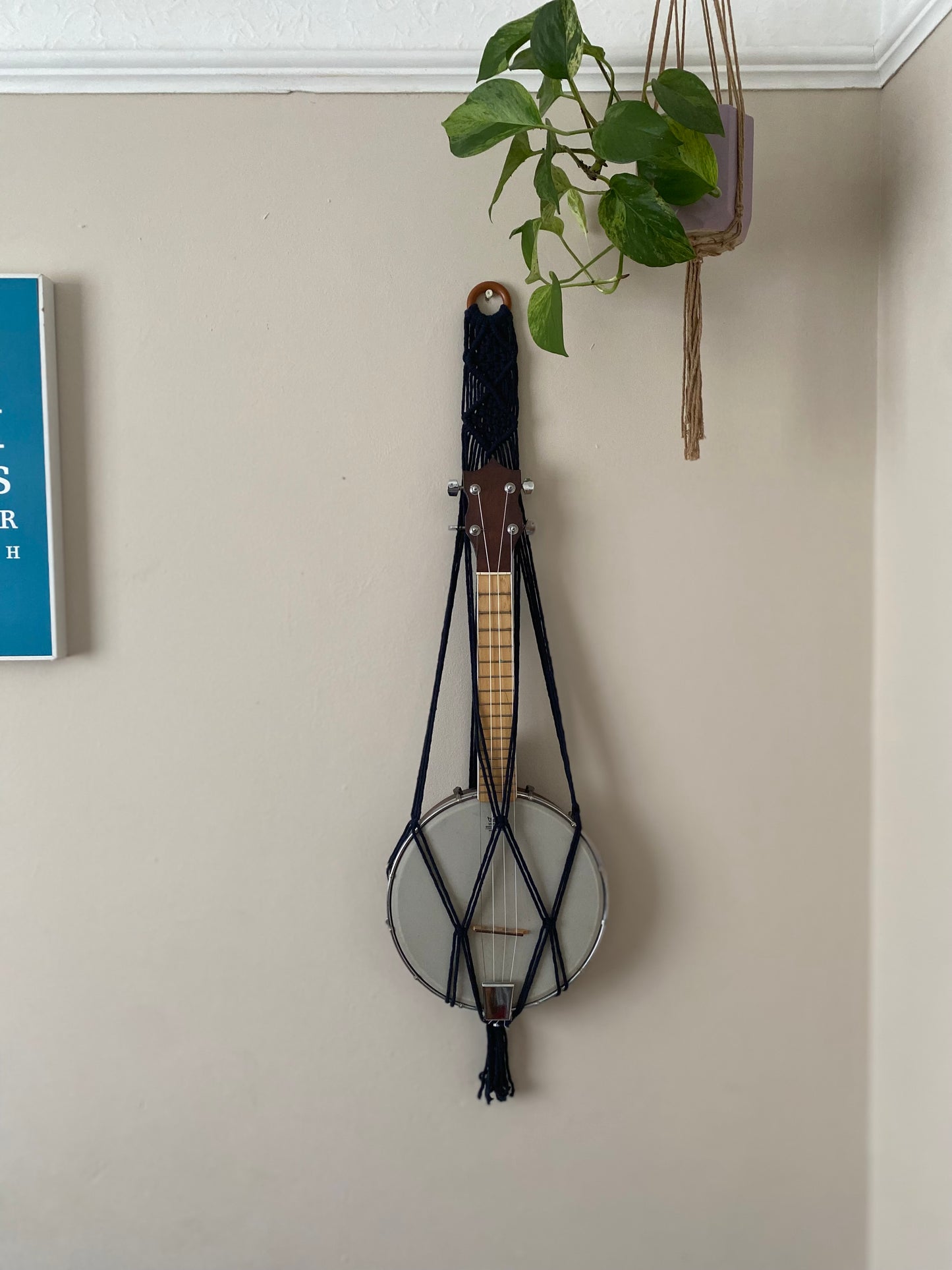 A banjolele instrument sits securely in a macrame wall mounted hanger, made with navy blue coloured yarn and a wooden hanging ring. Displayed against a beige painted wall, under a hanging houseplant.