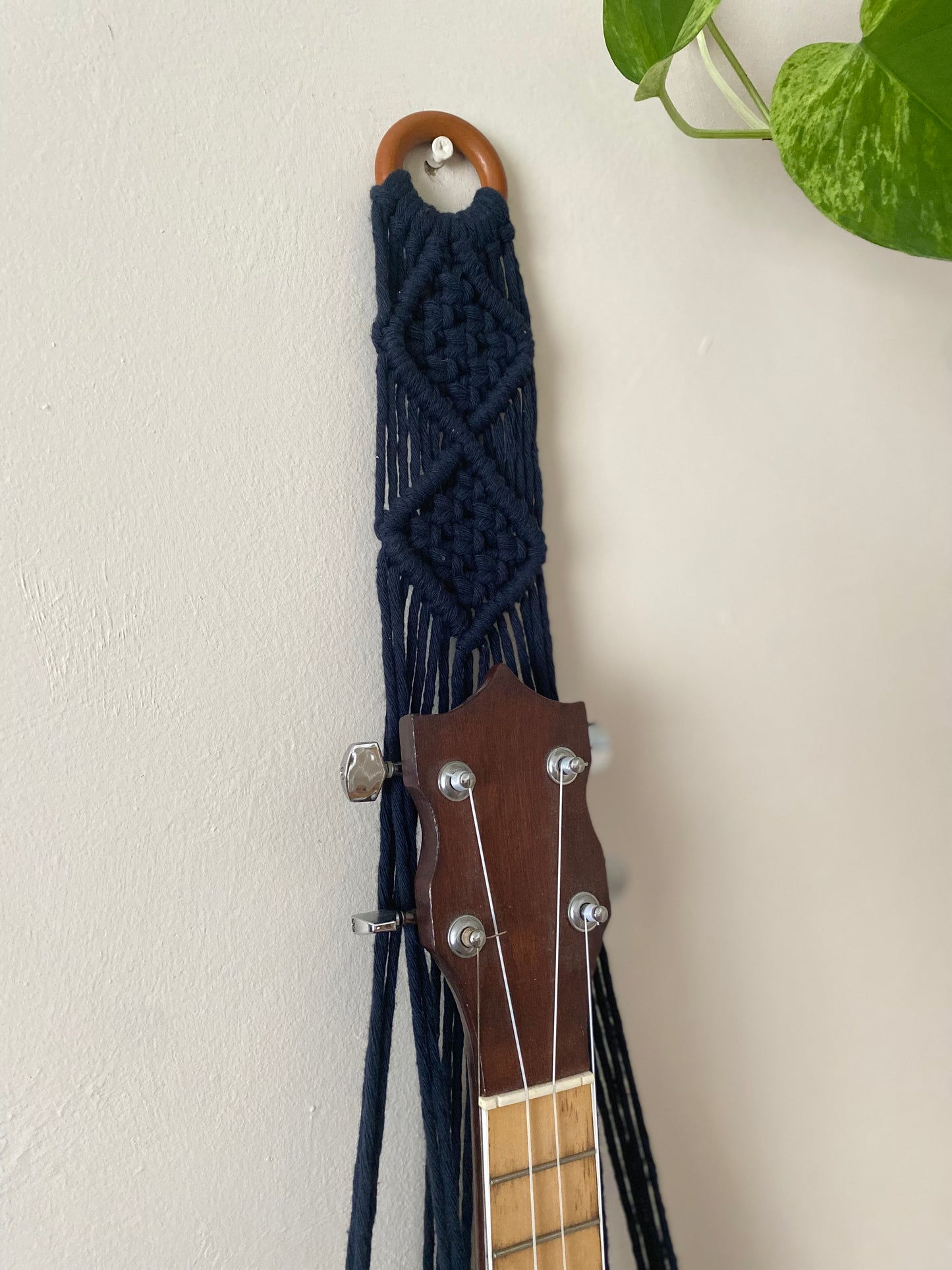 Close up image of the top section of a banjolele instrument in a macrame wall mounted hanger, made with navy blue coloured yarn and a wooden hanging ring.