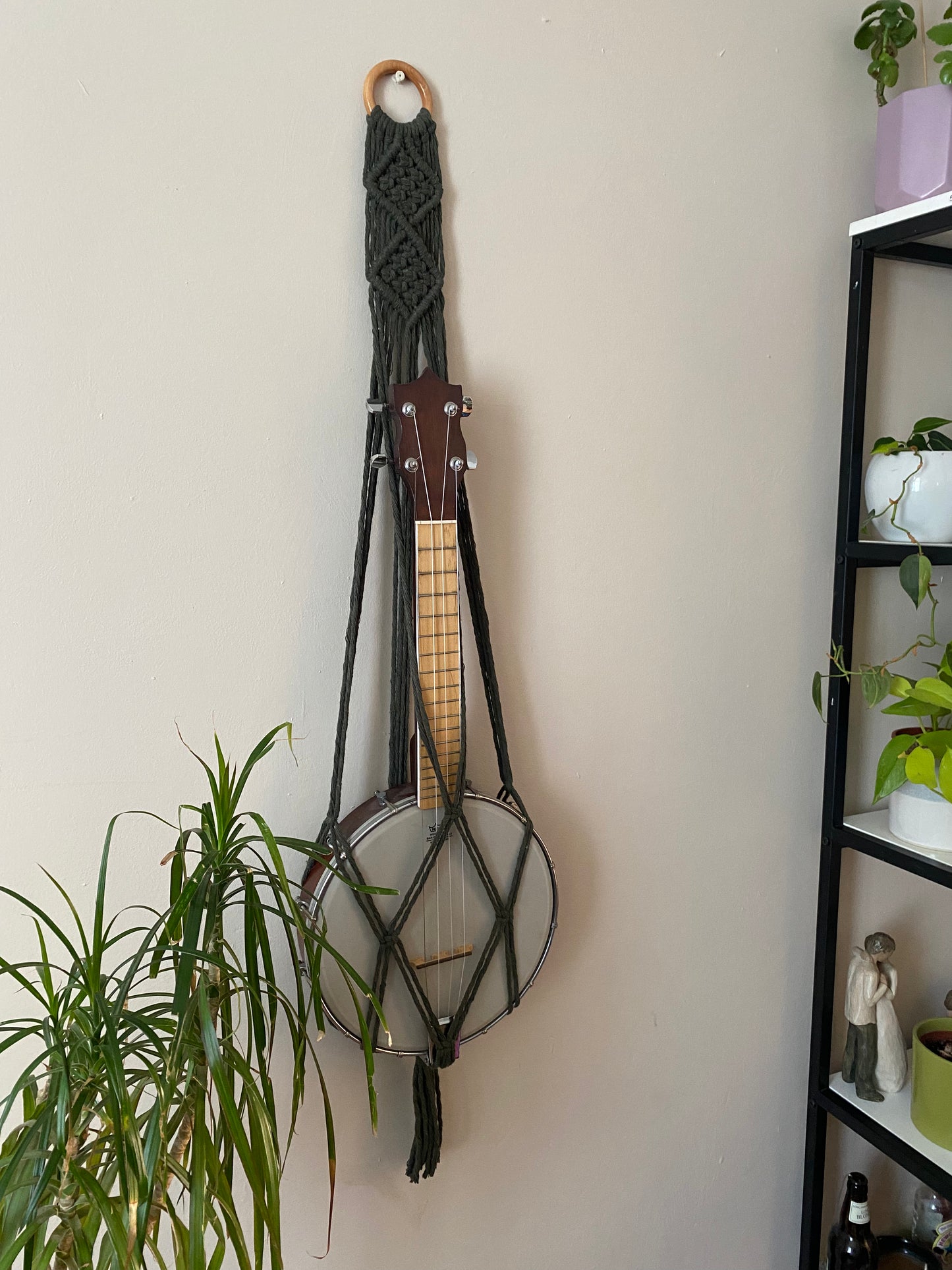 A banjolele instrument sits securely in a macrame wall mounted hanger, made with chunky olive green coloured yarn and a wooden hanging ring. Displayed against a beige painted wall, with various houseplants and a tall shelving unit nearby.