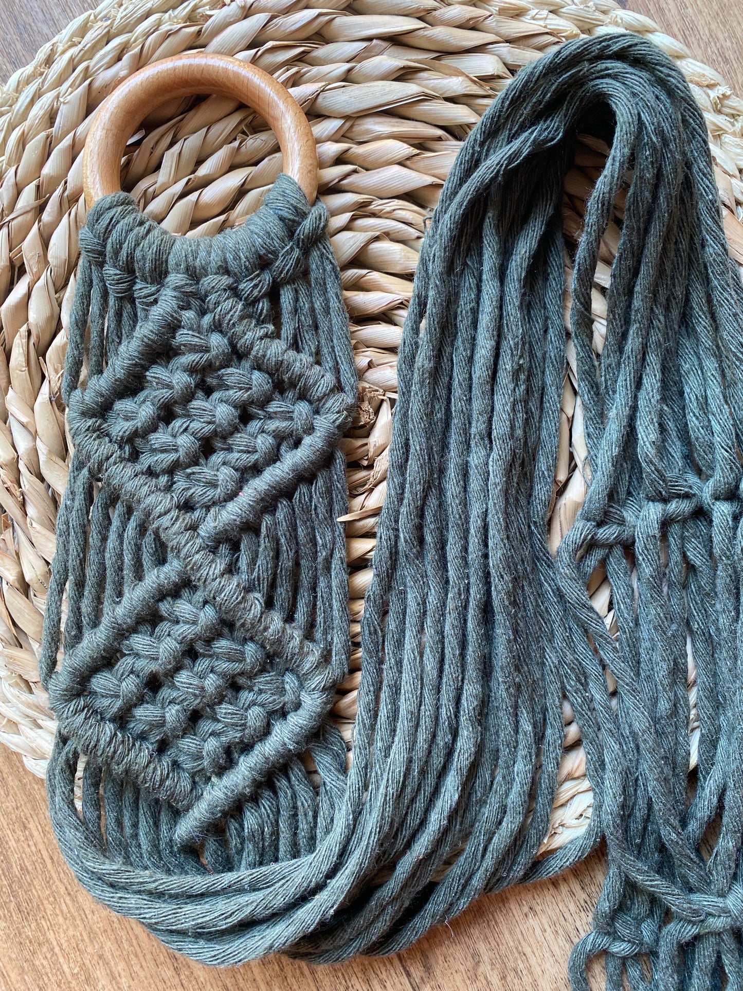 A handmade macrame instrument hanger made with chunky dark green yarn and a wooden ring, is folded neatly across a woven surface.