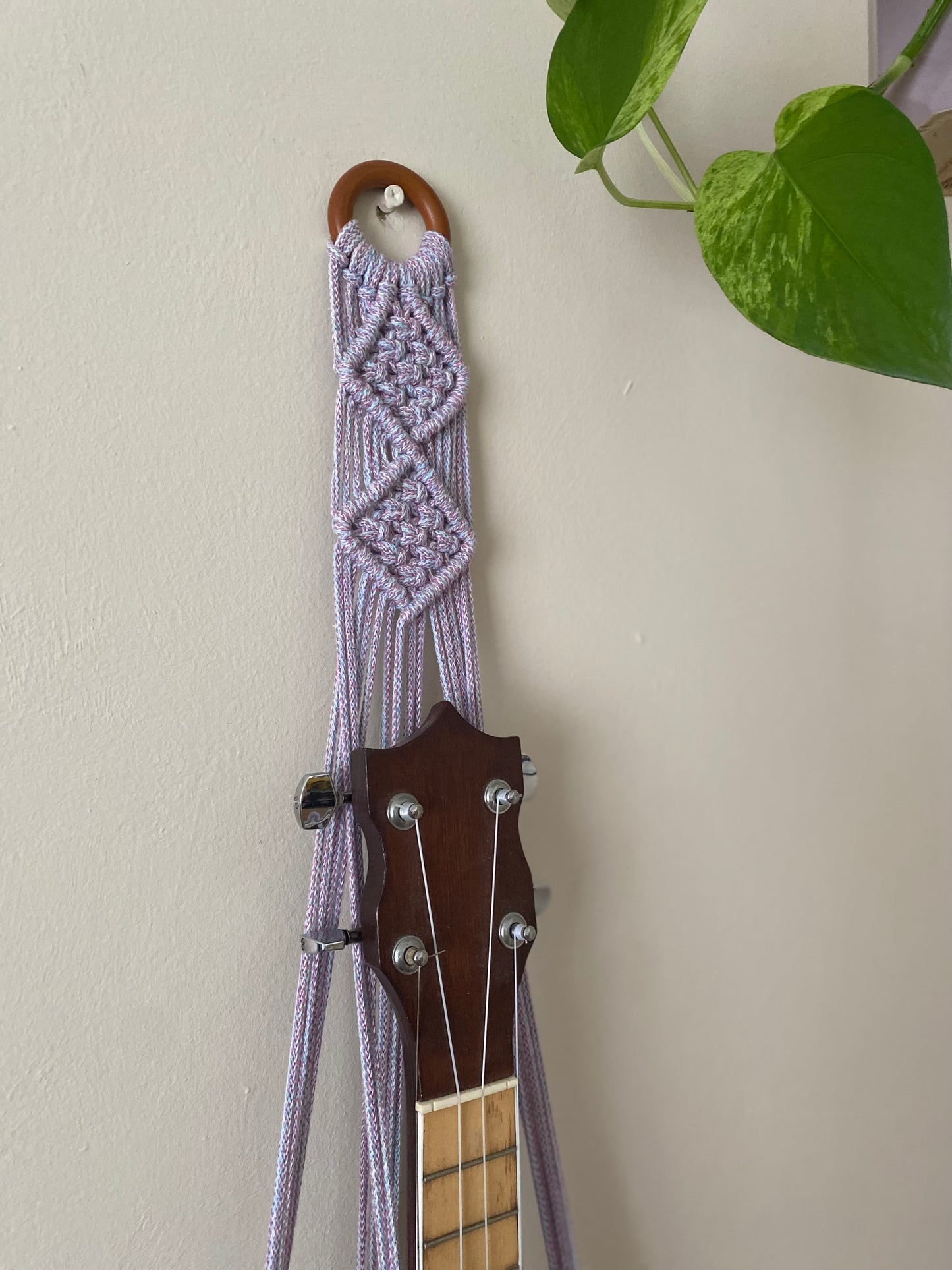 Close up of the top section of a banjolele instrument in a macrame wall mounted hanger, made with pastel coloured yarn and a wooden hanging ring.