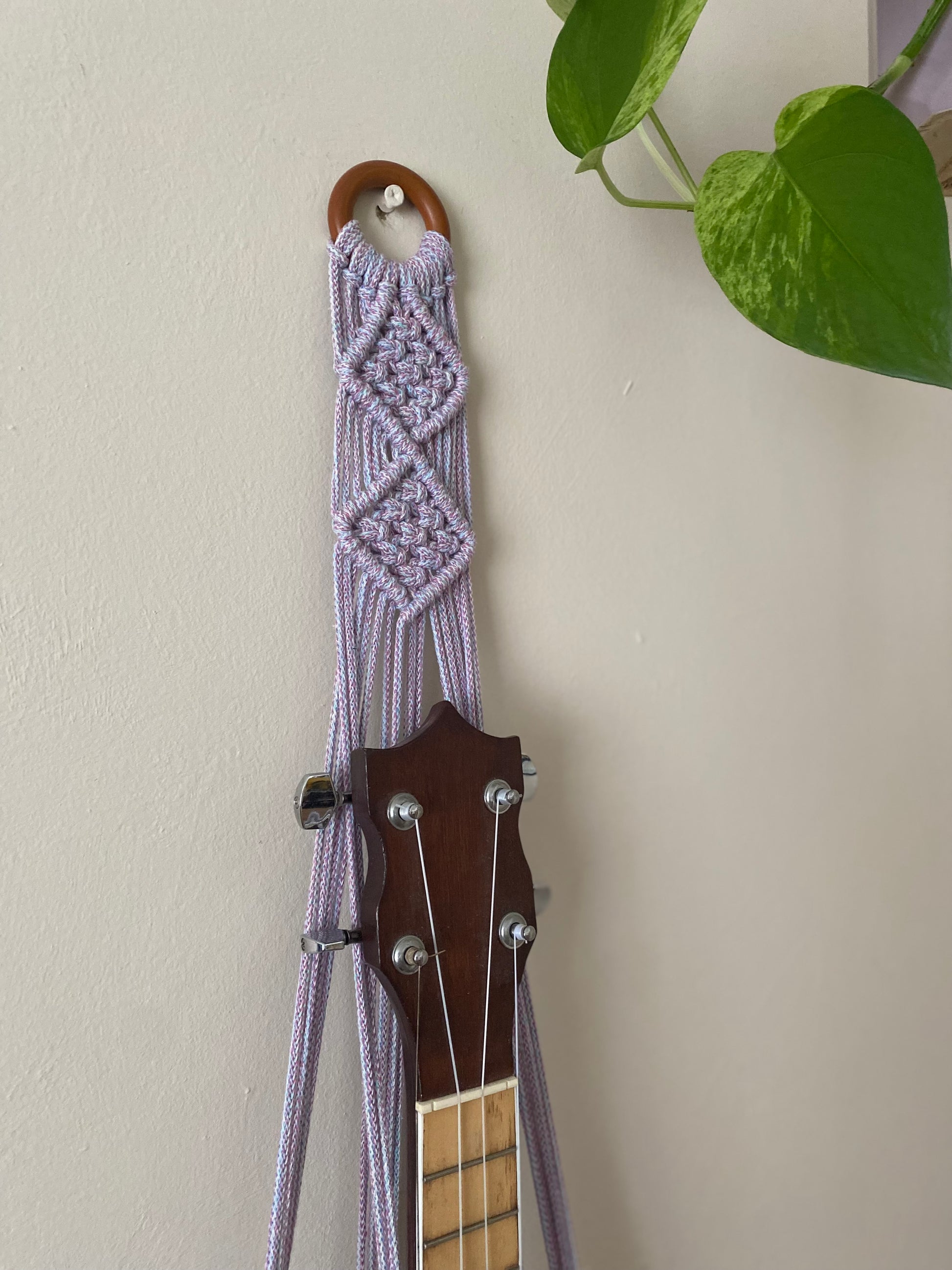 Close up of the top section of a banjolele instrument in a macrame wall mounted hanger, made with pastel coloured yarn and a wooden hanging ring.
