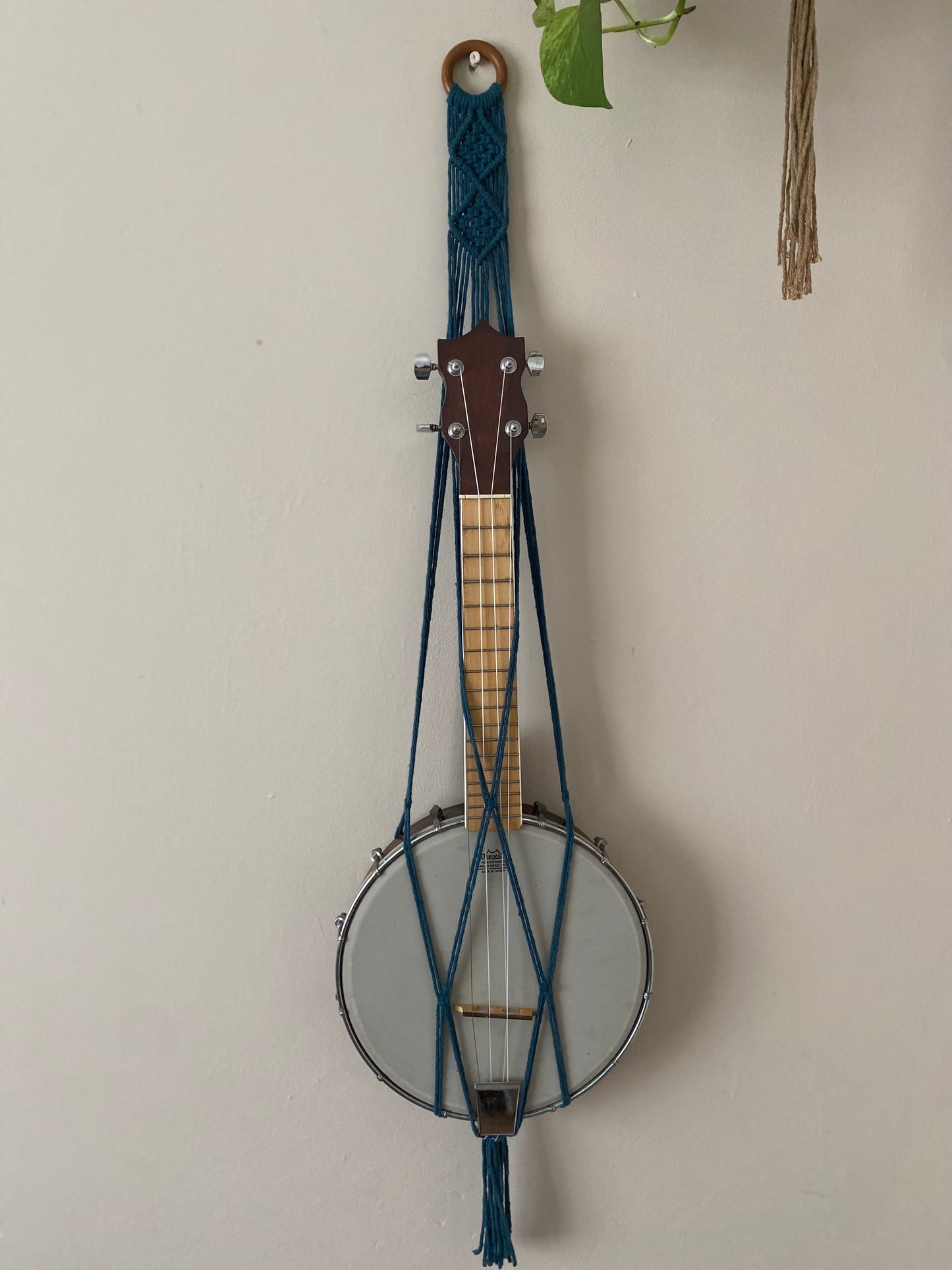 A banjolele instrument sits securely in a macrame wall mounted hanger, made with petrol blue coloured yarn and a wooden hanging ring. Displayed against a beige painted wall.
