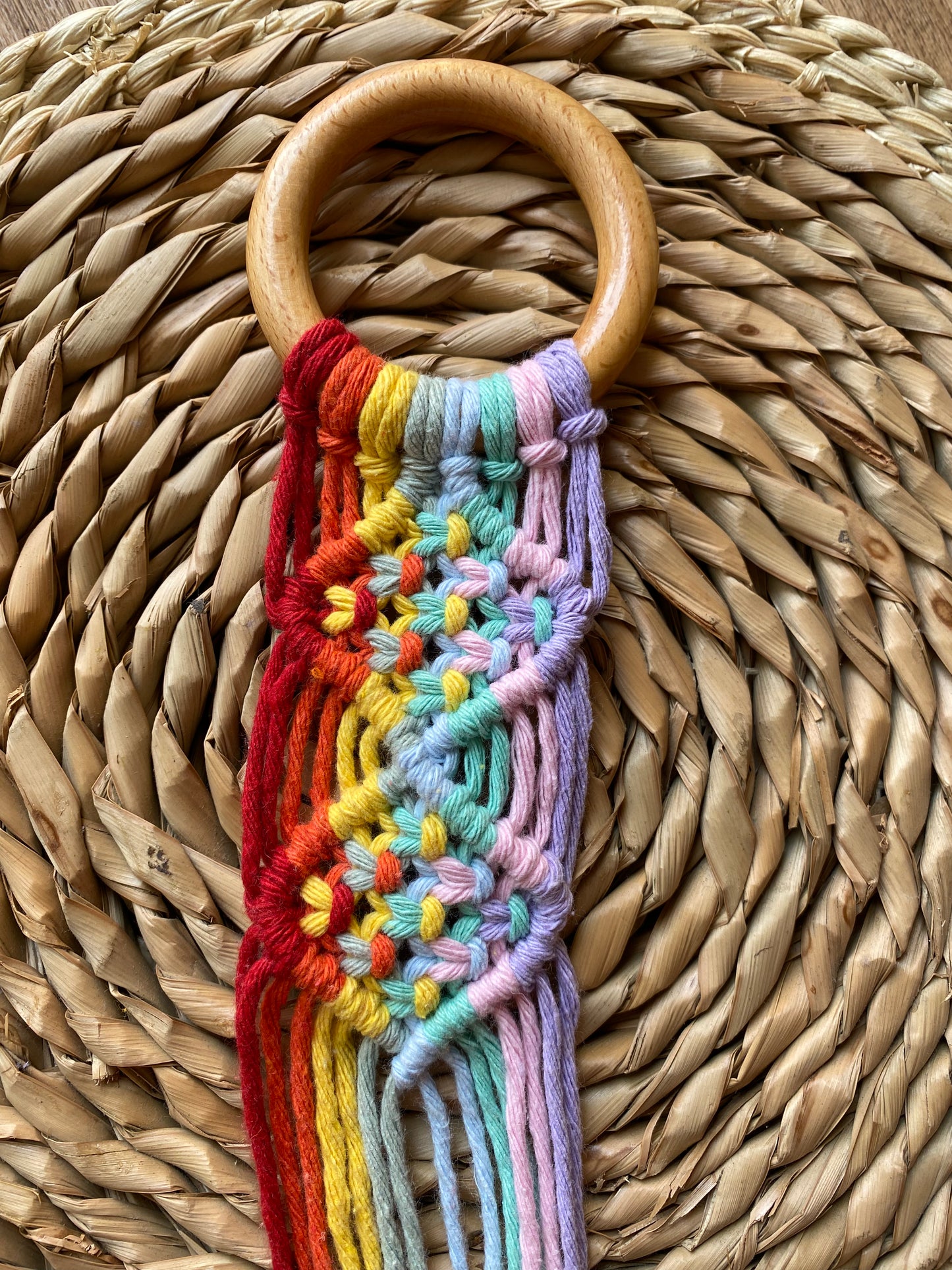 Macrame instrument hanger made with bright rainbow coloured yarn on a wooden ring with intricate knot pattern, laid on a woven wicker mat.