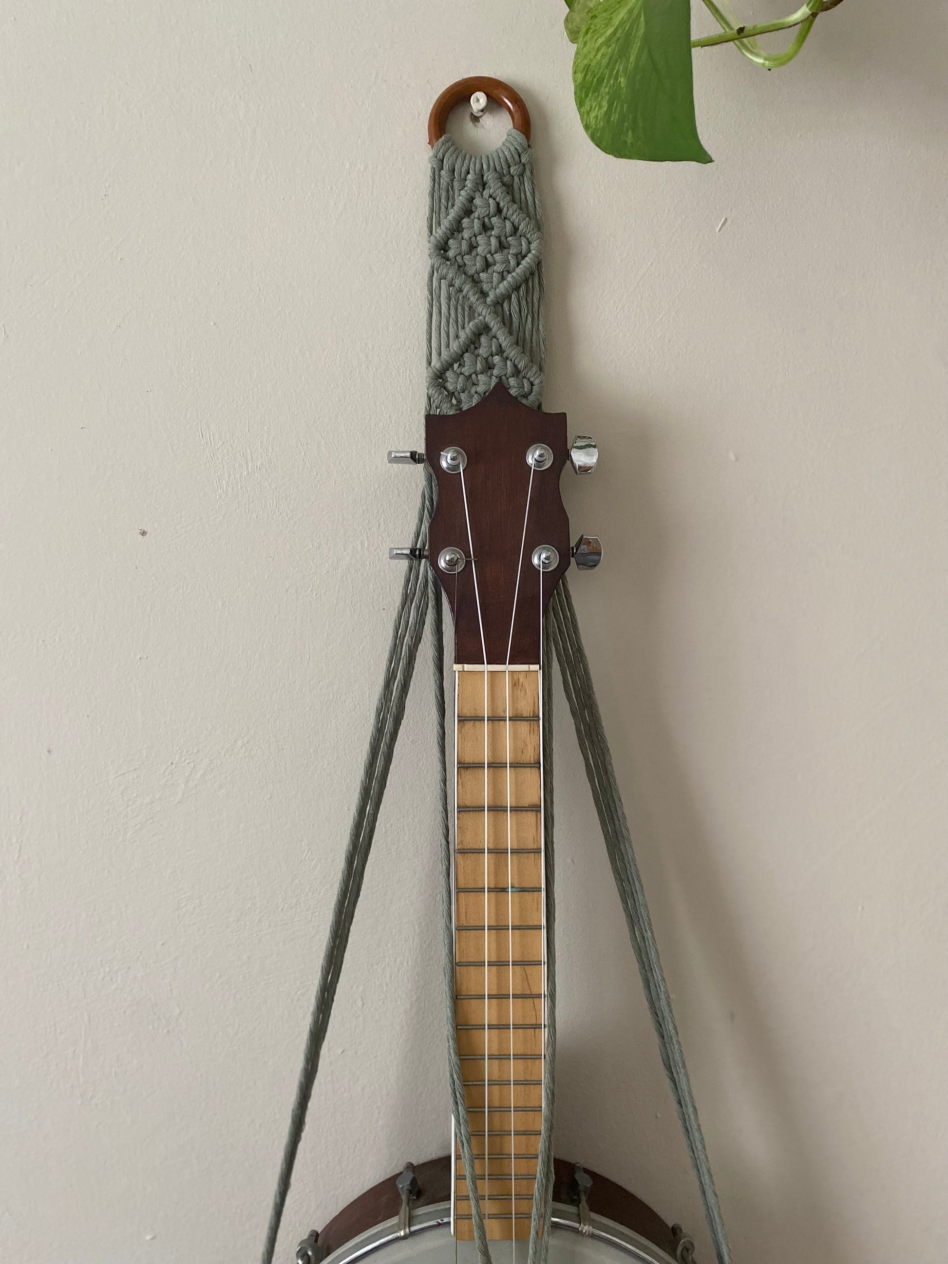 Close up image of the top section of a banjolele instrument in a macrame wall mounted hanger, made with sage green coloured yarn and a wooden hanging ring.