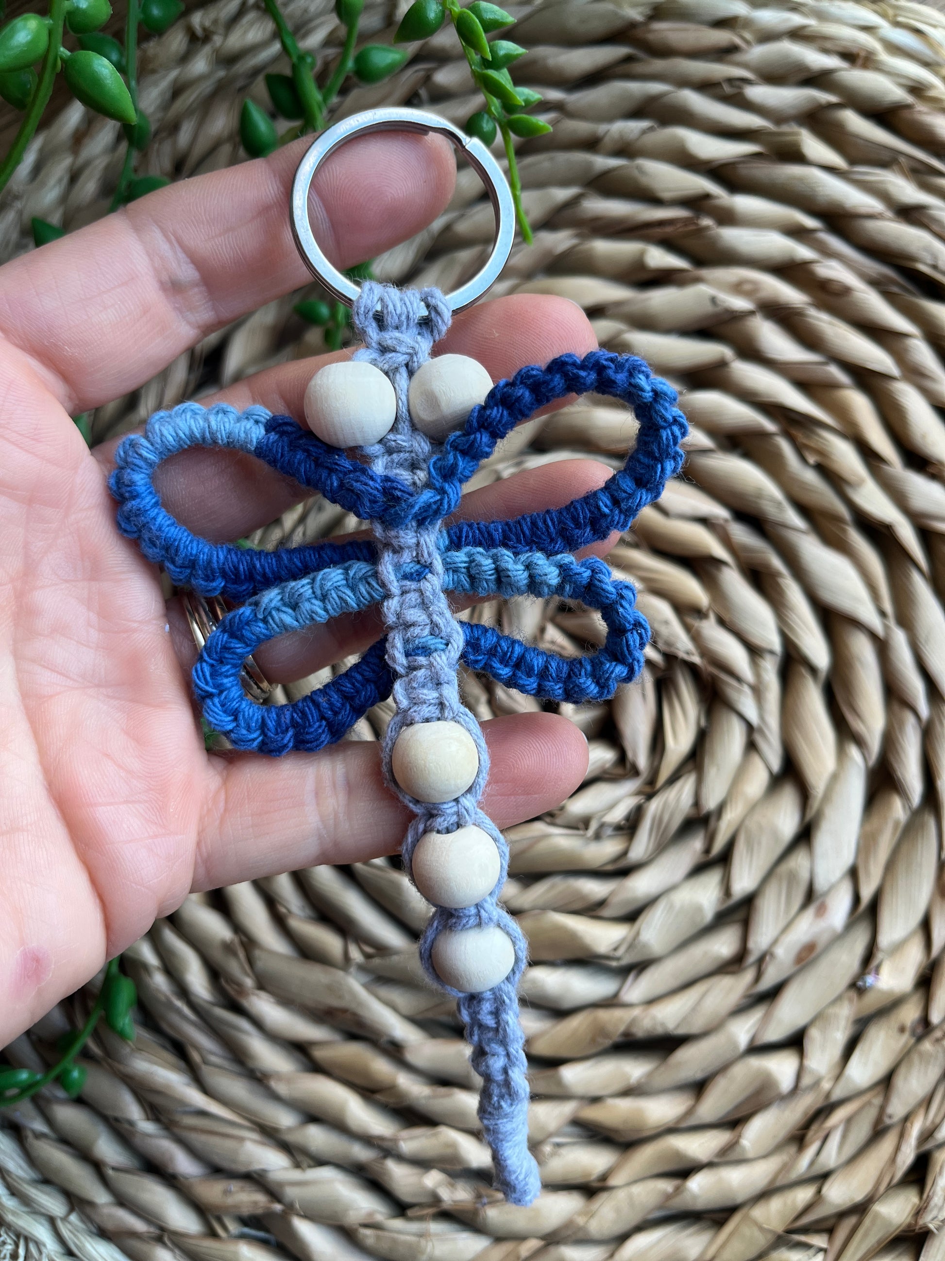 A hand holding a handmade macrame dragonfly keyring or bag charm, made using shades of blue recycled cotton, on a metal split ring, with small wooden beads.