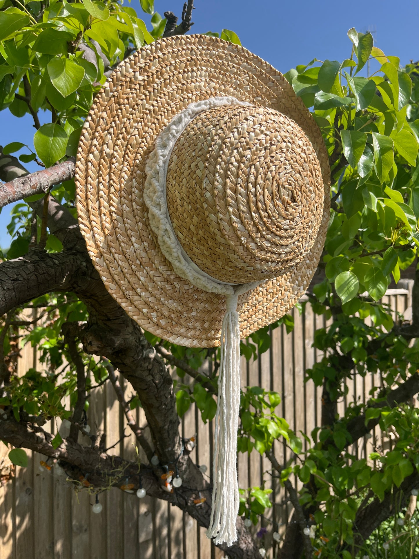 A wicker hat hangs from the branch of a pear tree, with a handmade boho style macrame hat band wrapped around the crown of the hat, with a long tassel.