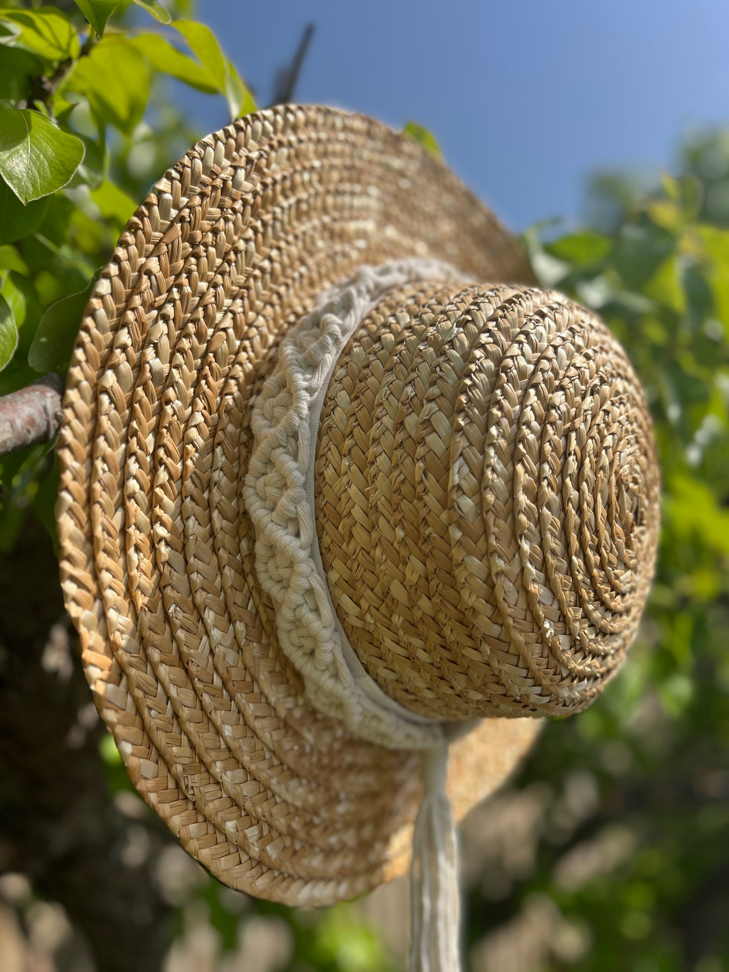 A wicker hat hangs from the branch of a pear tree, with a handmade boho style macrame hat band wrapped around the crown of the hat, with a long tassel.