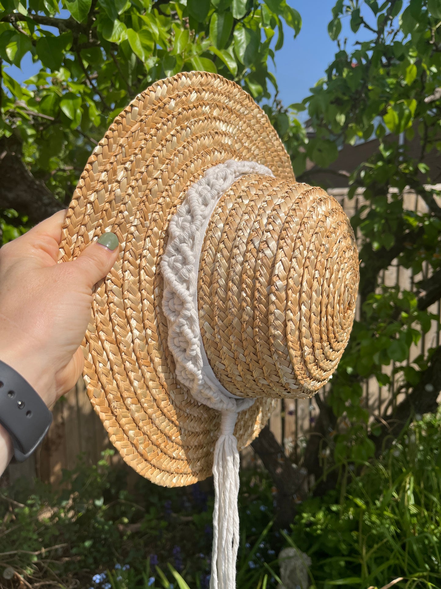 A wicker hat being held by the rim, outside in front of a pear tree, with a handmade boho style macrame hat band wrapped around the crown of the hat, with a long tassel.