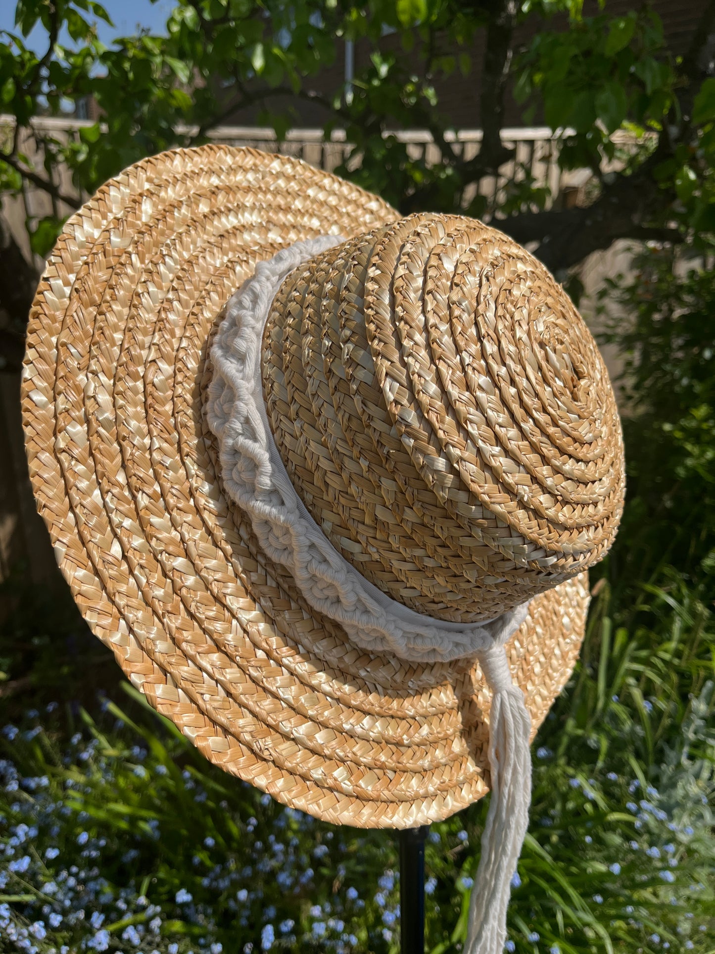 A wicker hat hangs in front of leafy green plants, outside, with a handmade boho style macrame hat band wrapped around the crown of the hat, with a long tassel.