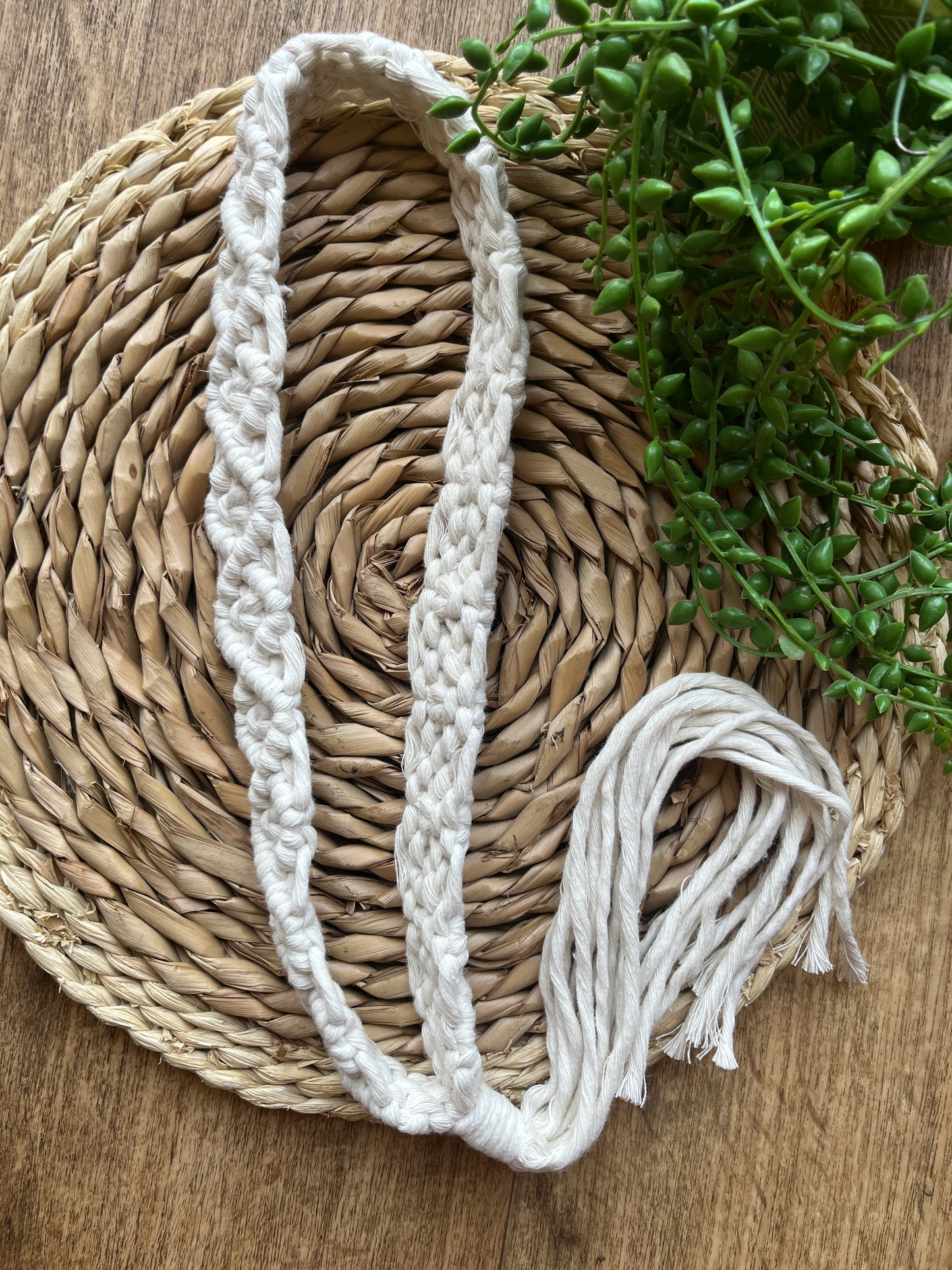 A handmade boho style macrame hat band with a long tassel, made with recycled cotton yarn, laid on a round wicker surface next to some green plant vines.