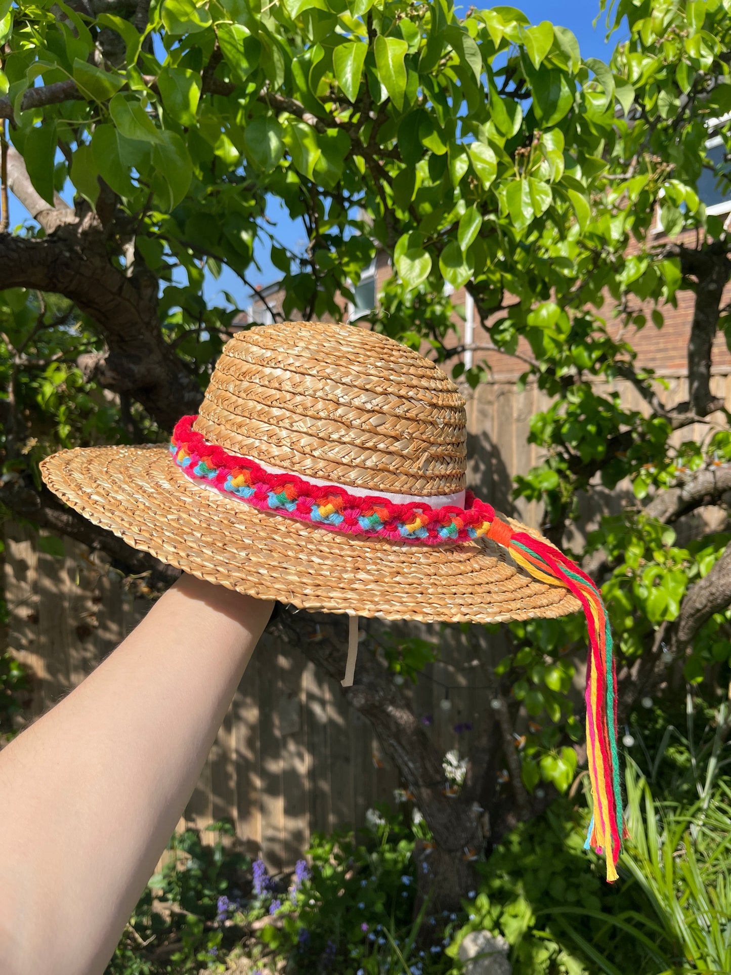 A wicker hat held outside in front of a pear tree, with a handmade boho style macrame hat band wrapped around the crown of the hat, with a long tassel. The hat band is made with rainbow colours of recycled cotton.