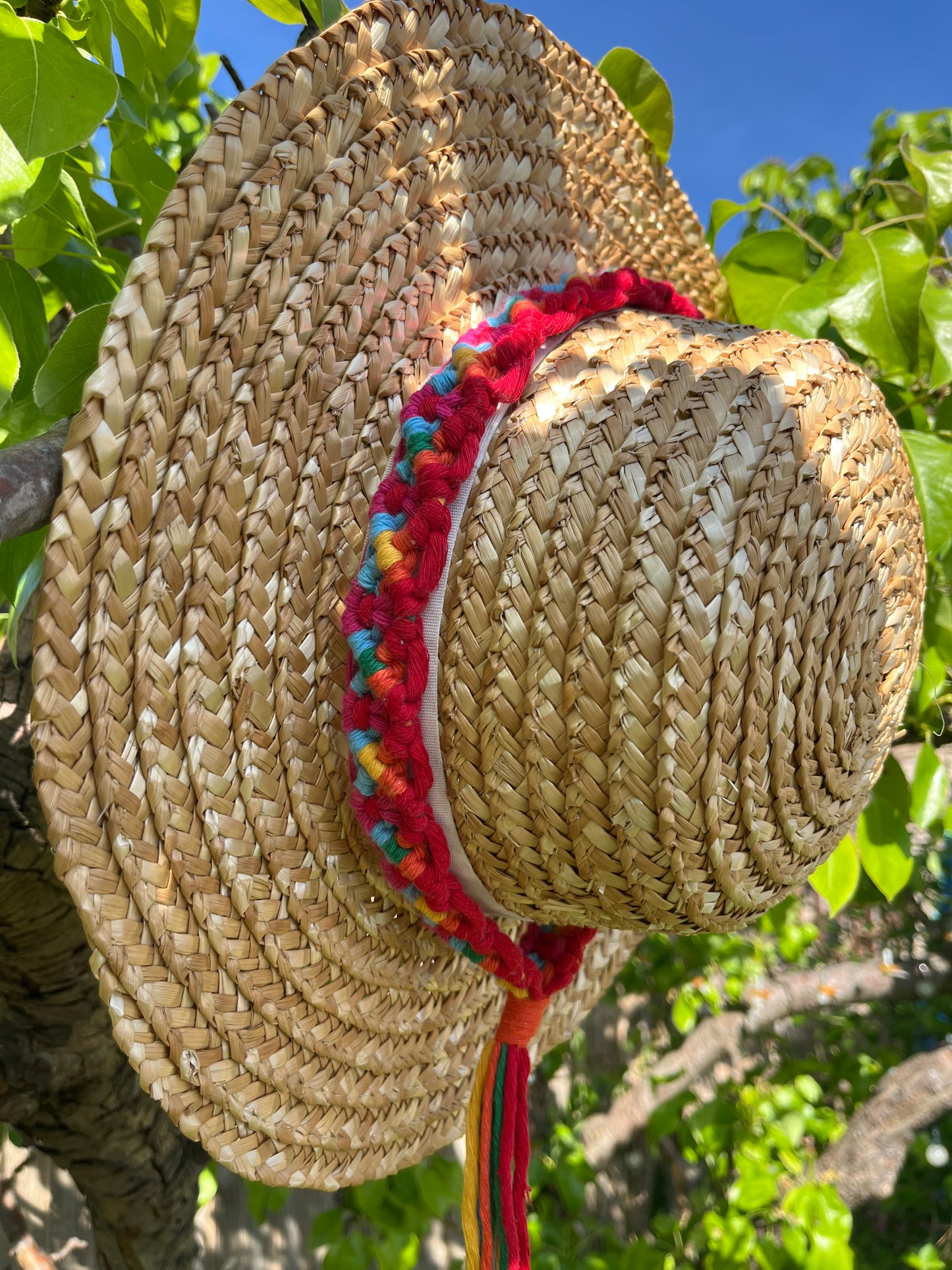 A wicker hat hangs outside in front of a pear tree, with a handmade boho style macrame hat band wrapped around the crown of the hat, with a long tassel. The hat band is made with rainbow colours of recycled cotton.