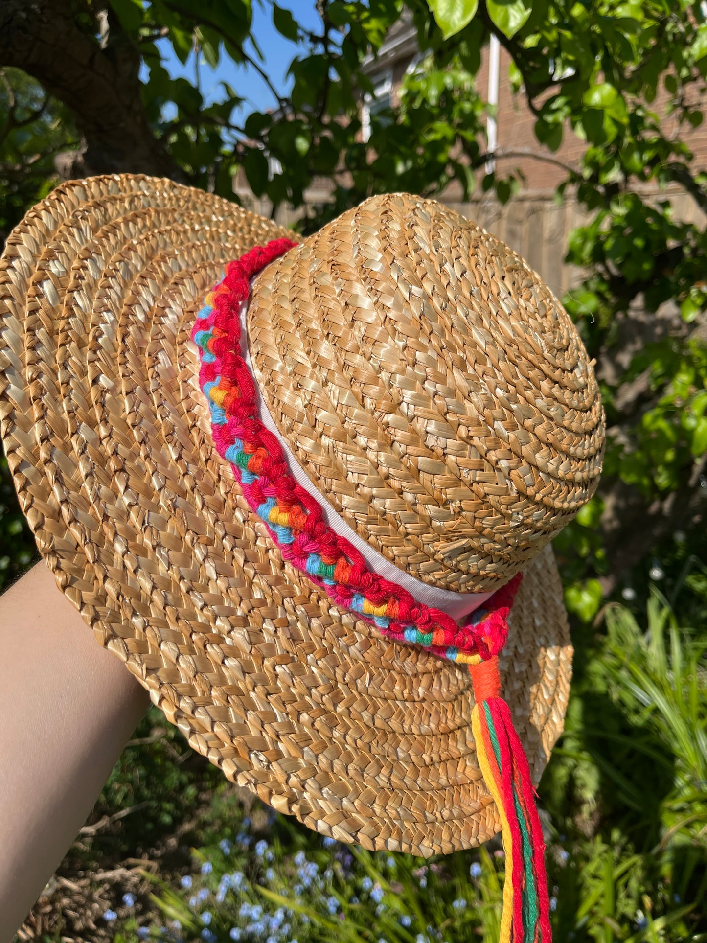 A wicker hat held outside in front of a pear tree, with a handmade boho style macrame hat band wrapped around the crown of the hat, with a long tassel. The hat band is made with rainbow colours of recycled cotton.