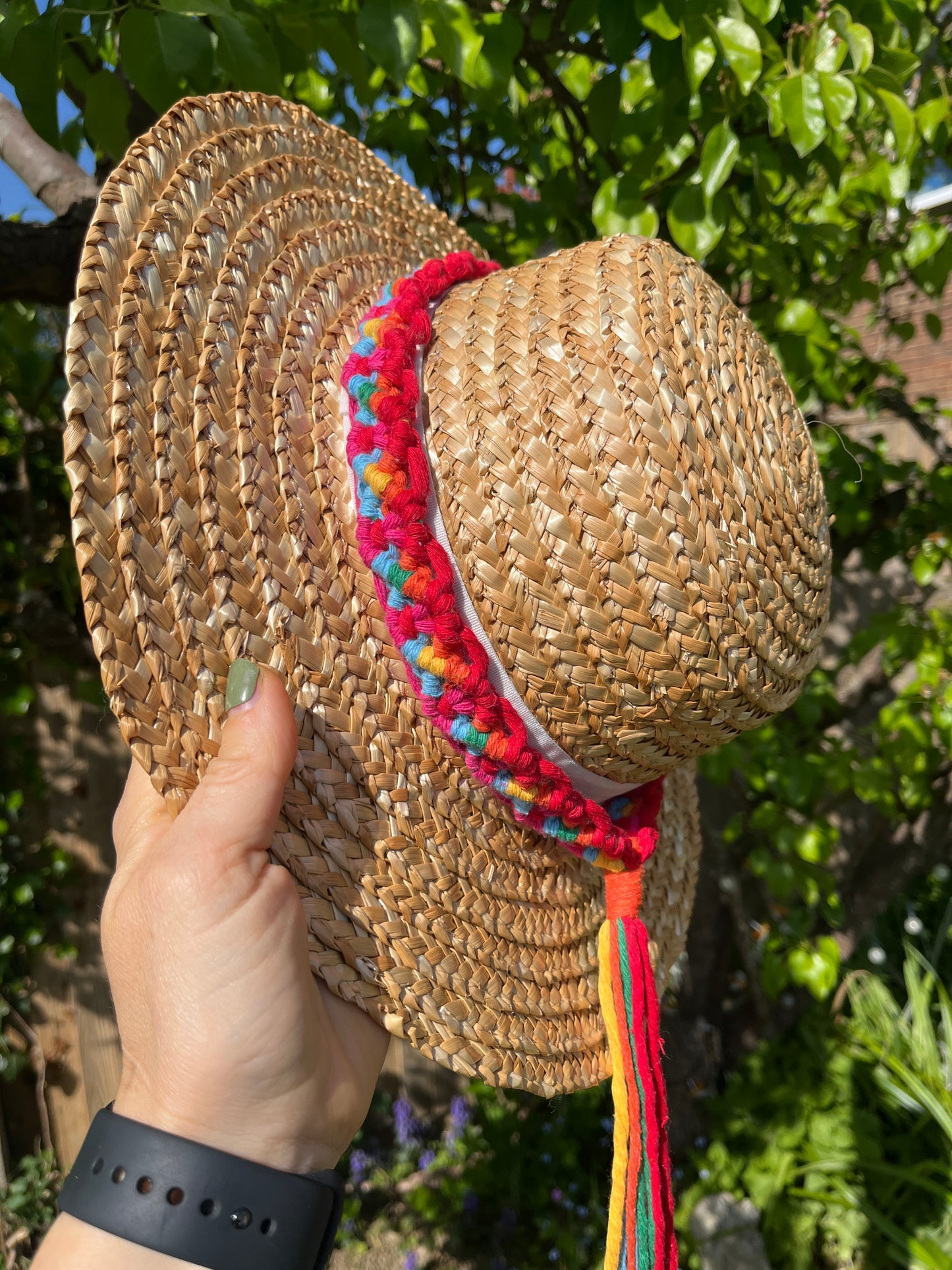 A wicker hat held outside by the rim, in front of a pear tree, with a handmade boho style macrame hat band wrapped around the crown of the hat, with a long tassel. The hat band is made with rainbow colours of recycled cotton.