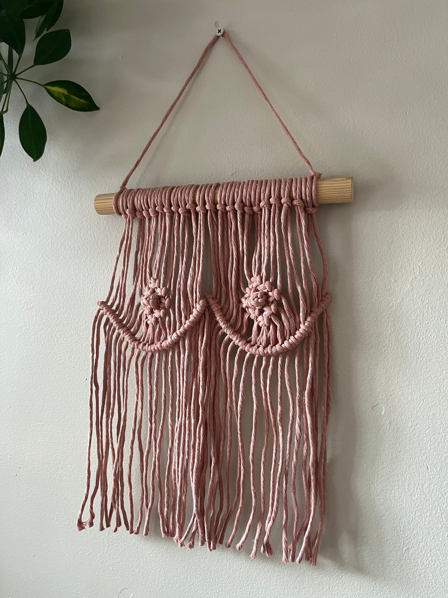A macrame wall hanging representing boobs, made with blush pink yarn on a wooden dowel, hanging on a beige wall.