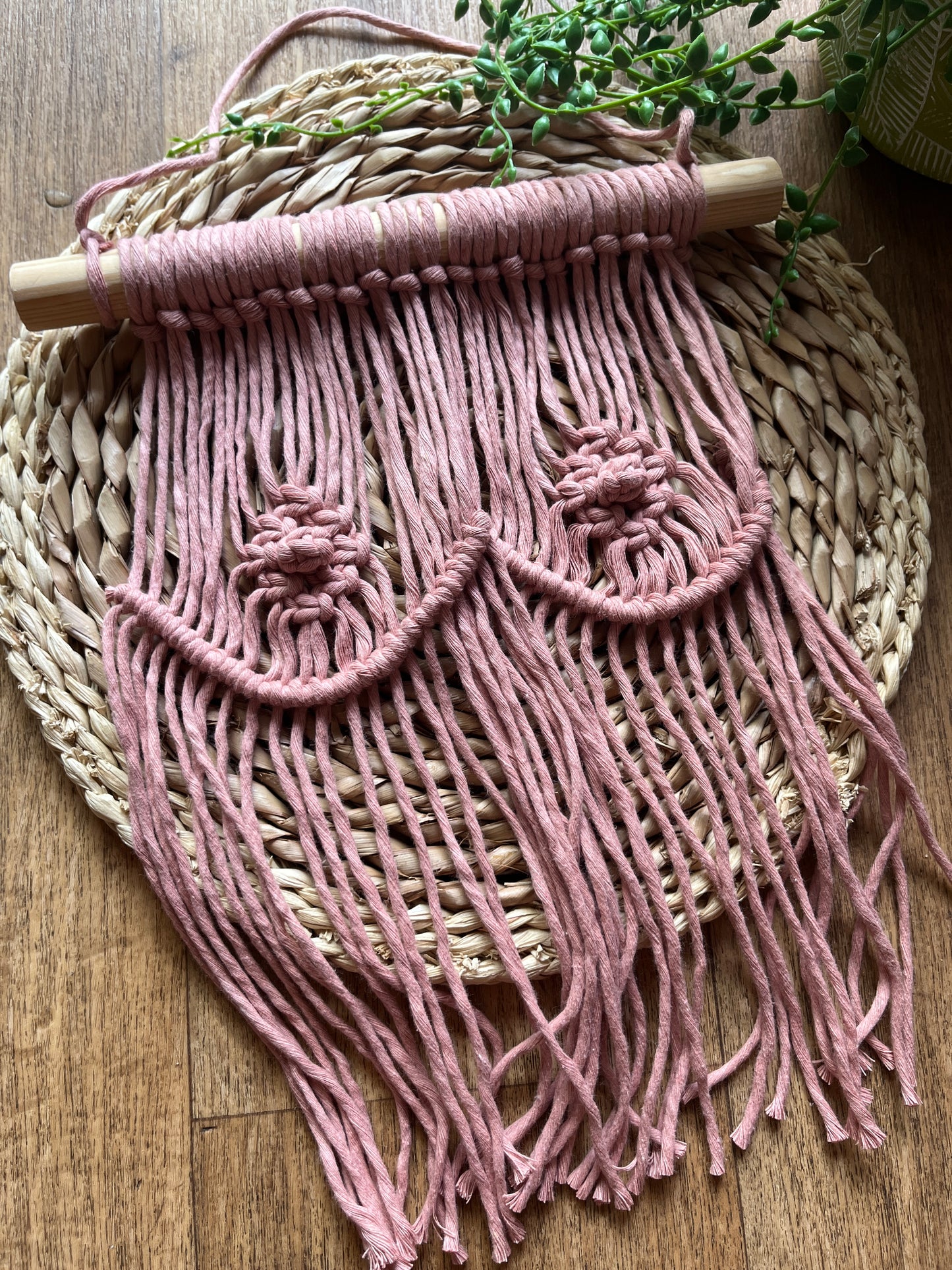 A macrame wall hanging representing boobs, made with blush pink yarn on a wooden dowel, laid across a round wicker mat.