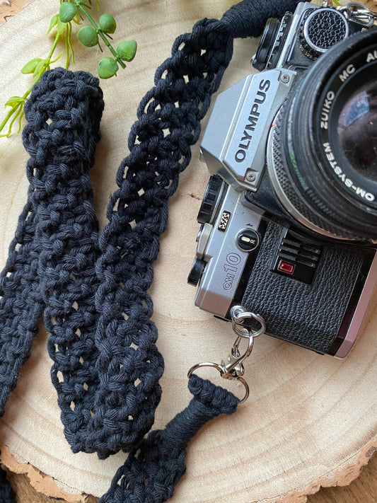 A black Olympus vintage camera lays on a round wooden surface, with a handmade macrame knotted camera strap attached to it with metal clasps. The camera strap is made with black yarn.
