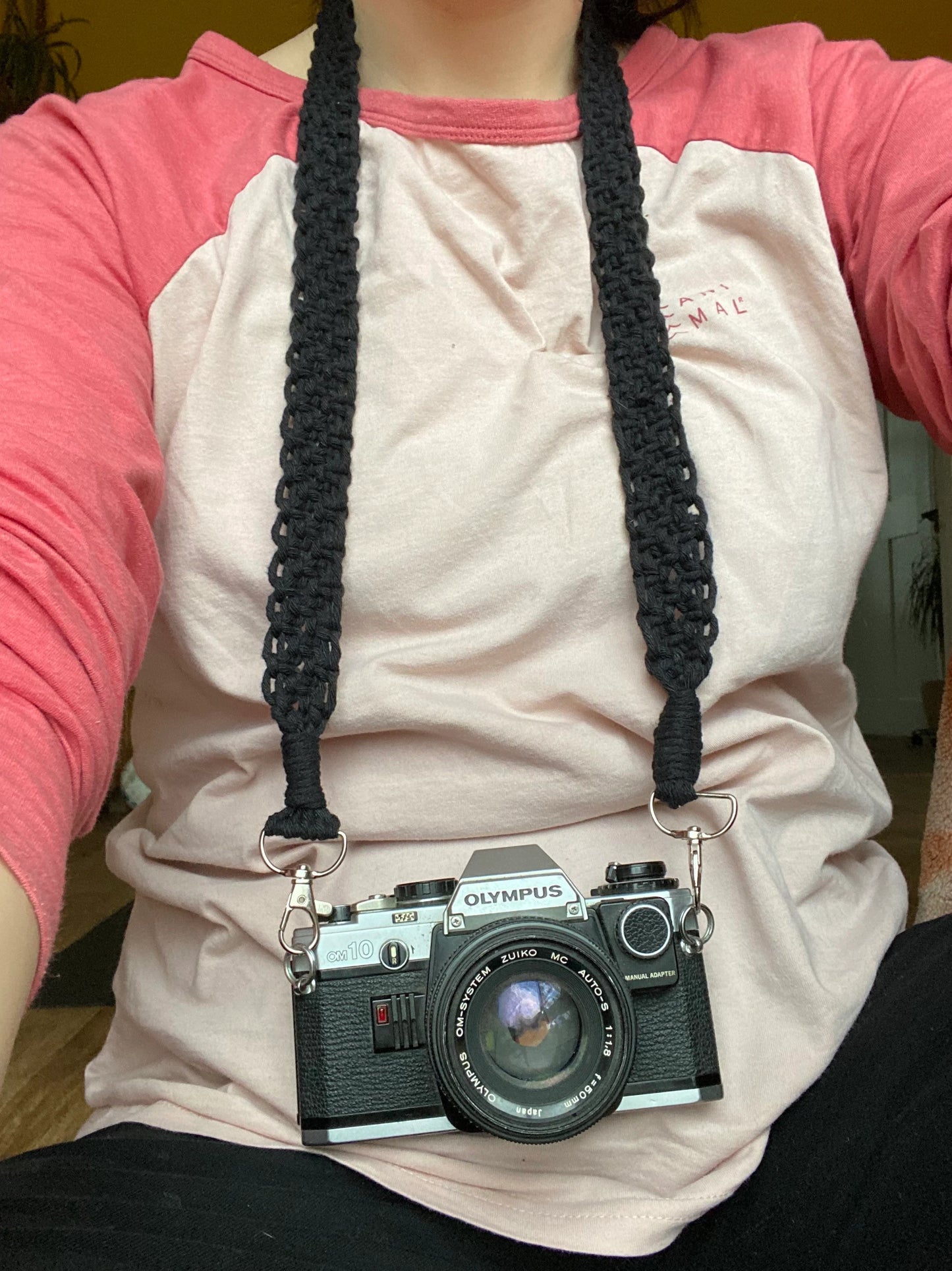 A woman sits wearing a pink t shirt, with a black Olympus vintage camera held around her neck on a handmade macrame knotted camera strap. The camera strap is made with black yarn and has metal clasps on each end..
