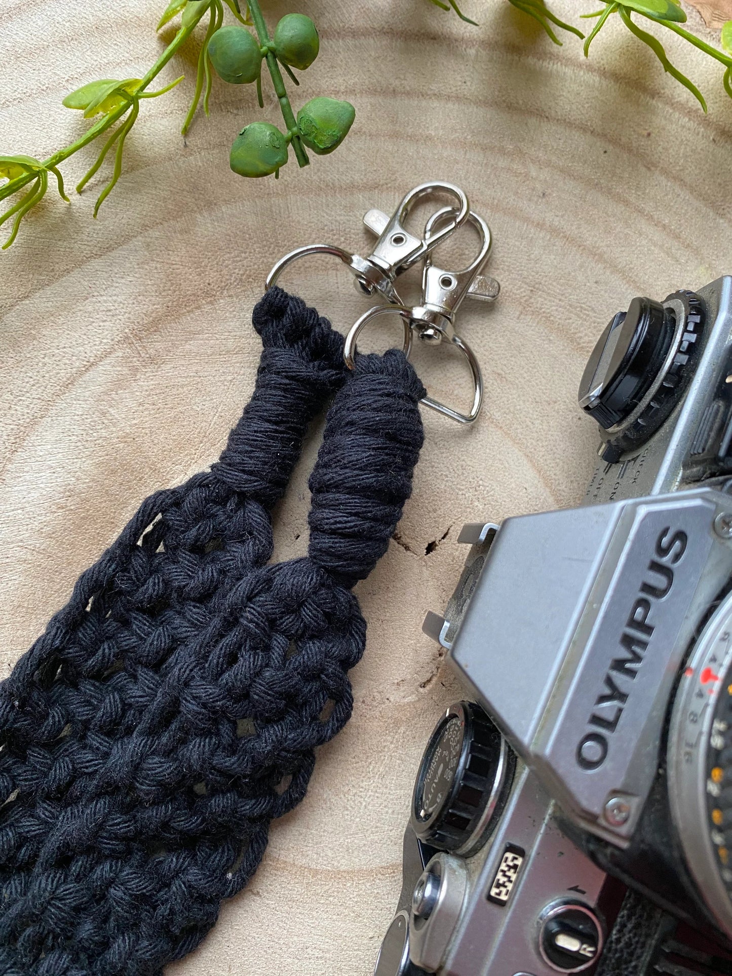 A black Olympus vintage camera lays on a round wooden surface, with a handmade macrame knotted camera strap next to it. The camera strap is made with black yarn and has metal clasps on each end.