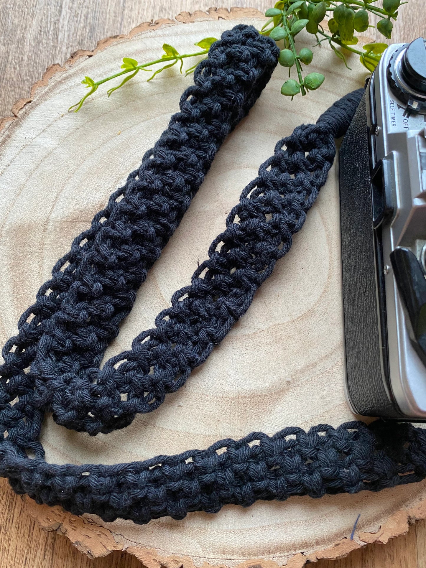 A black Olympus vintage camera sits on a round wooden surface, with a handmade macrame knotted camera strap attached to it with metal clasps. The camera strap is made with black yarn.