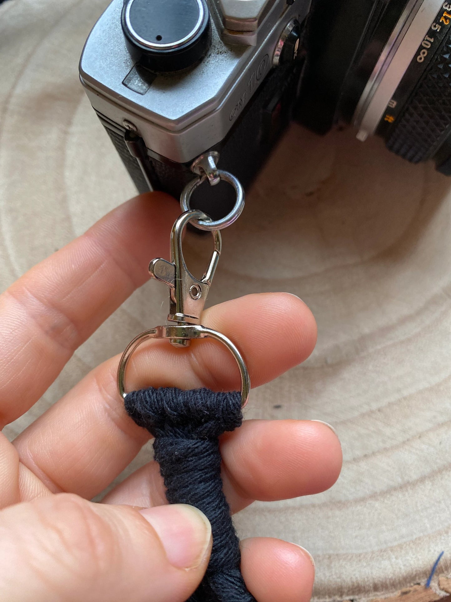 A black Olympus vintage camera sits on a round wooden surface, a hand is holding the metal clasp on the end of a handmade macrame knotted camera strap, where it attaches to the camera.