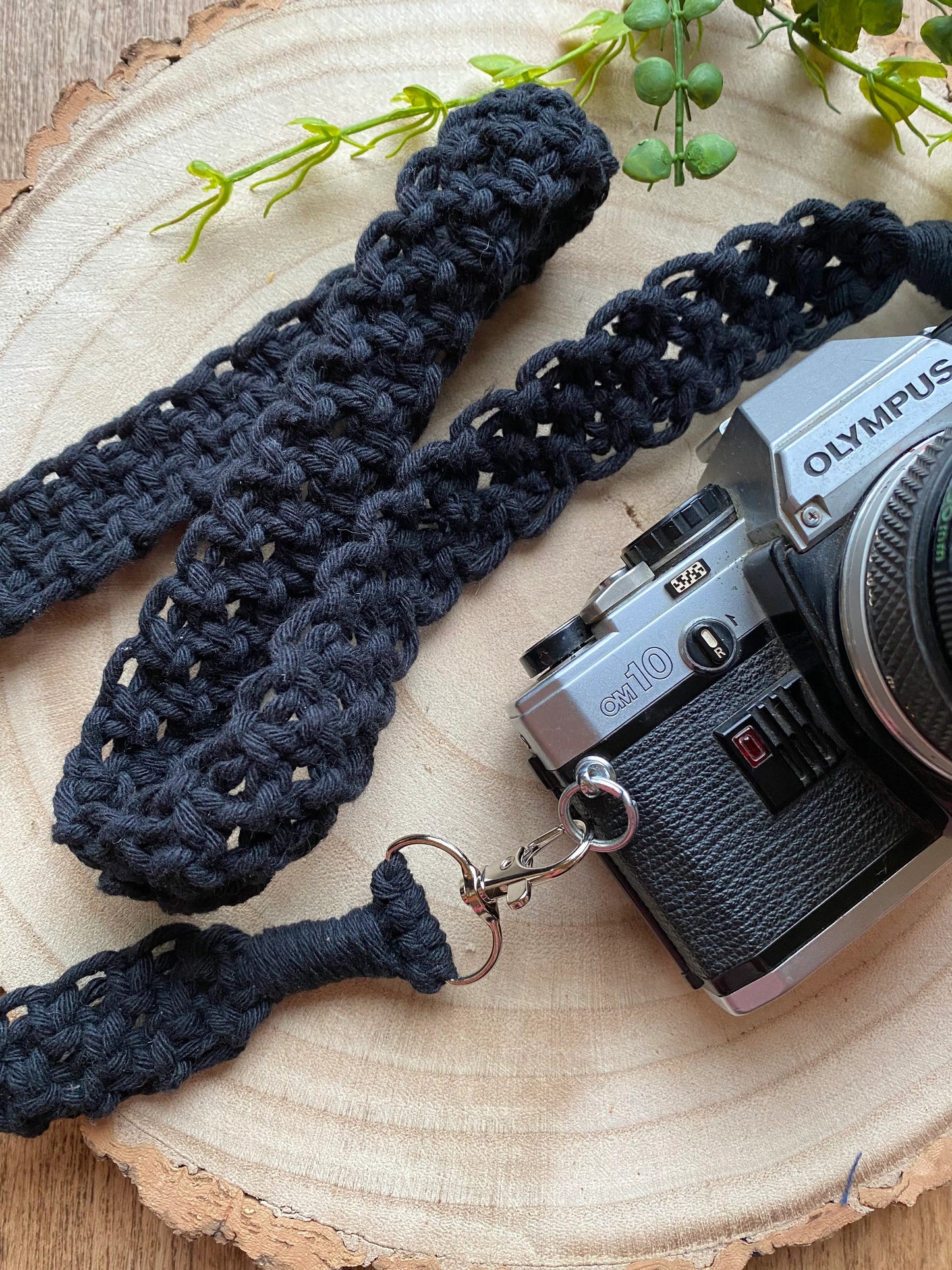 A top down view of a black Olympus vintage camera on a round wooden surface, with a handmade macrame knotted camera strap attached to it with metal clasps.