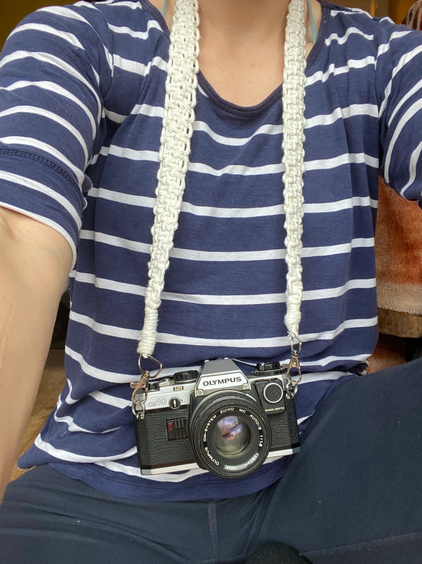 A woman sits wearing a blue and white striped top, with a black Olympus vintage camera held around her neck with a handmade macrame knotted camera strap attached to it with metal clasps. The camera strap is made with natural coloured yarn.